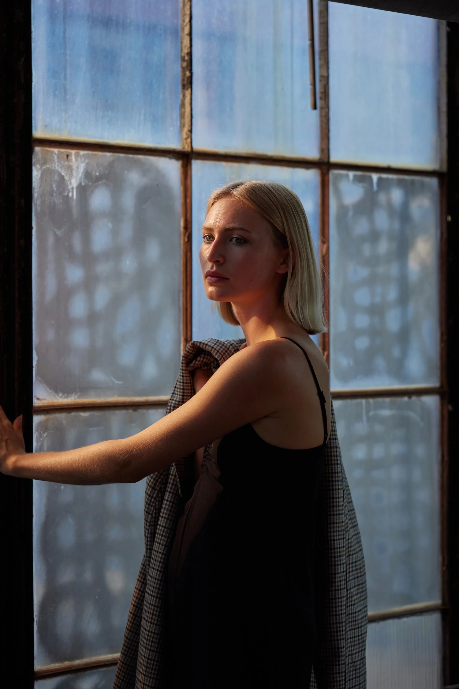 A young woman standing in front of a large industrial-style window, with natural light illuminating her face and shoulder. She is wearing a black spaghetti strap top and holding a checkered jacket over her shoulder.