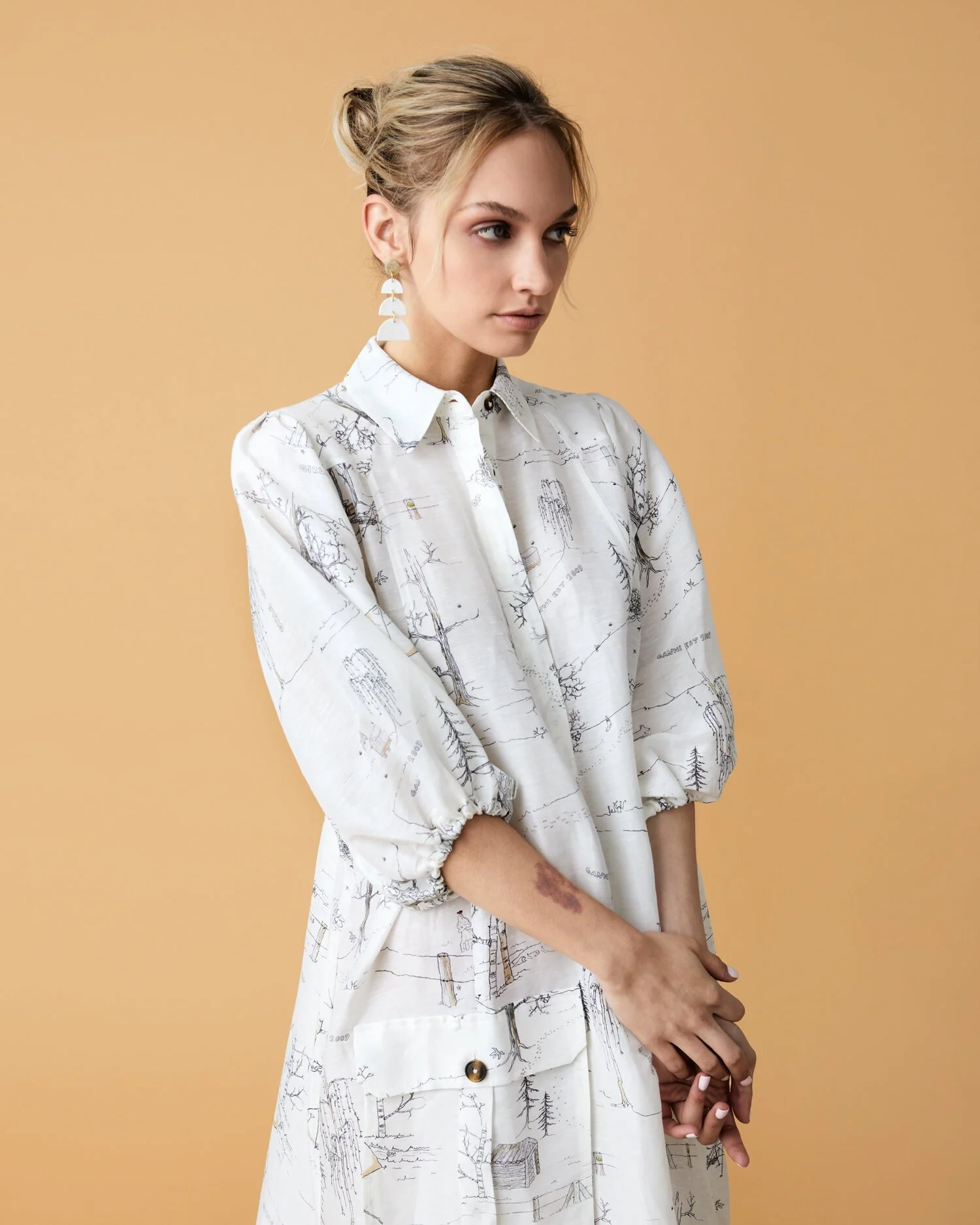 A young woman with blonde hair styled in an updo, wearing a white dress with a black line drawing pattern of trees and nature scenes, standing against a beige background.