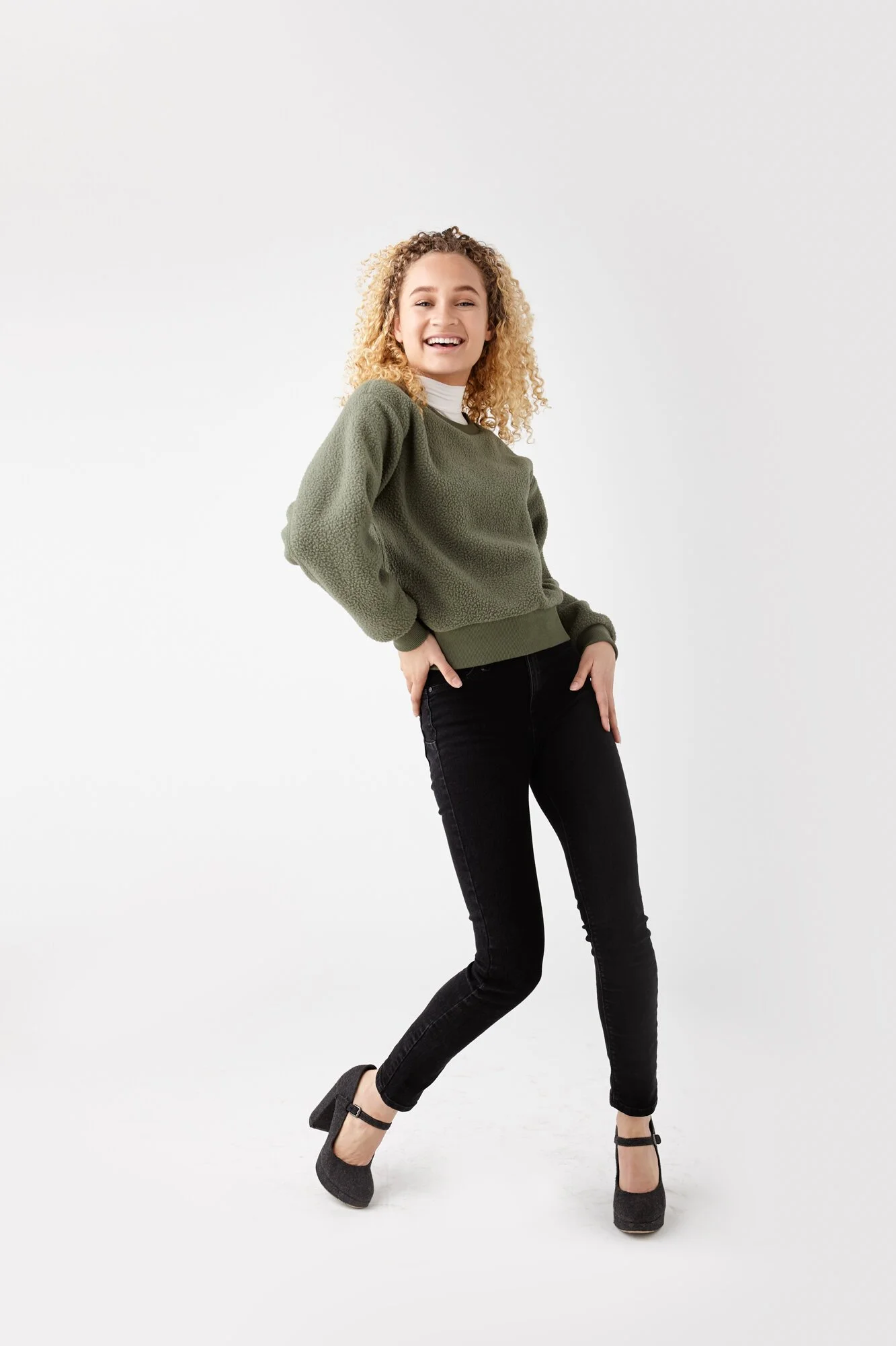 A young woman with curly blonde hair smiling and posing in casual clothing: green sweater, white shirt, black jeans, and gray heels, standing against a plain white background.