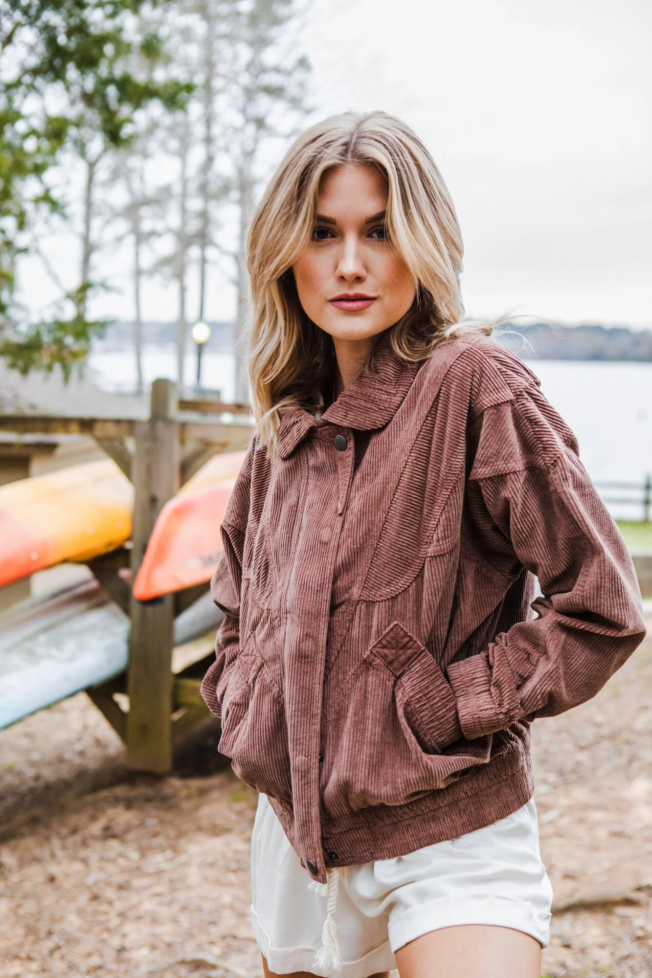 A woman standing outdoors on a cloudy day near a lake, wearing a brown corduroy jacket and white shorts, with kayaks in the background.