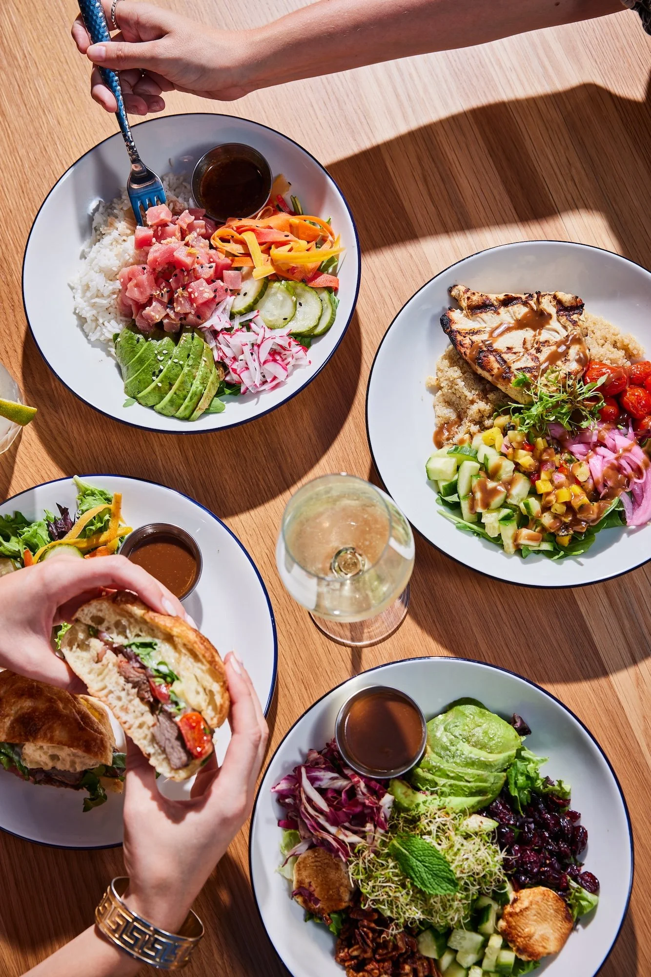 A top-down view of a table with three plates of colorful salads and grilled chicken, a sandwich, and a glass of white wine, with hands reaching for food.