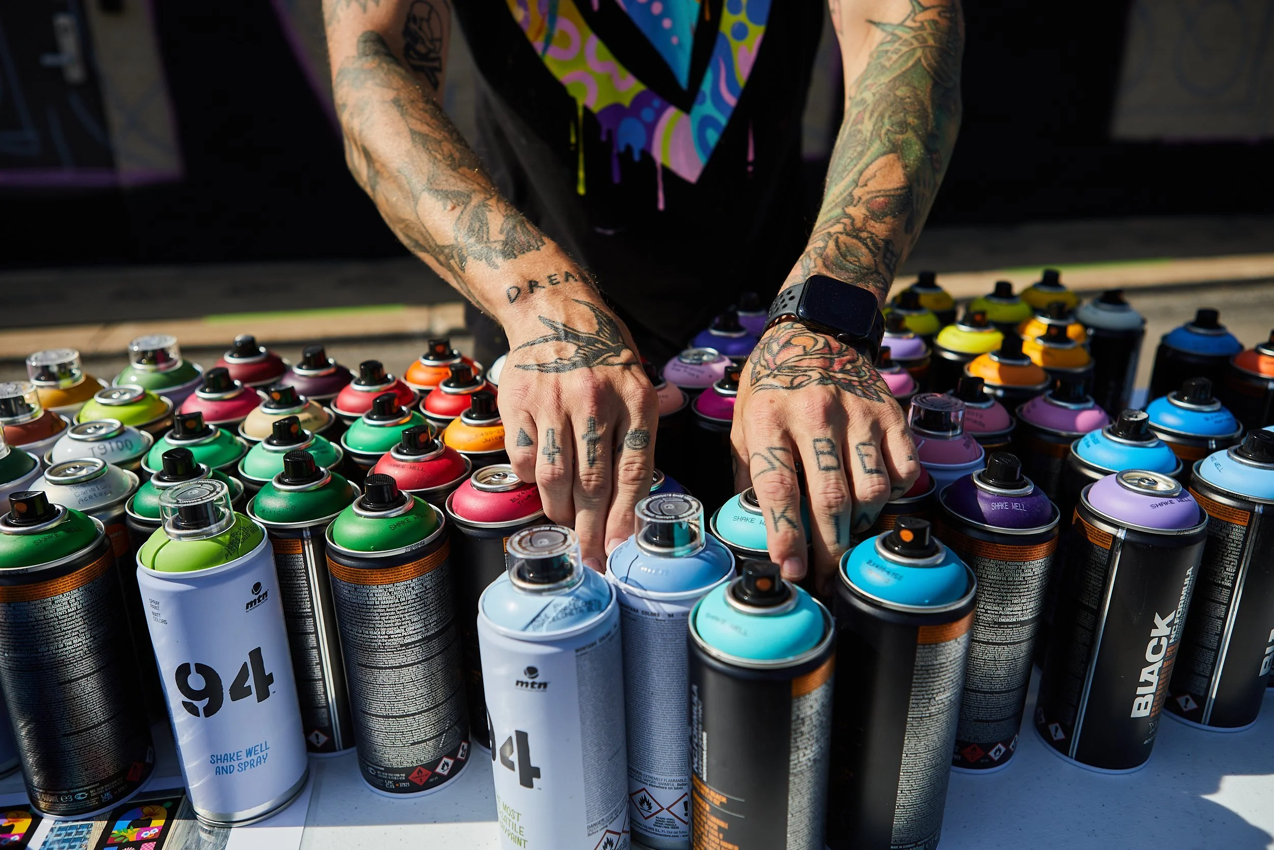 Person with tattoos standing at table of colorful spray paint cans, arranging the cans. The person's hands are positioned over the cans, and they are wearing a black shirt with a colorful, stylized 'Superman' logo. Tattoos are visible on their arms a