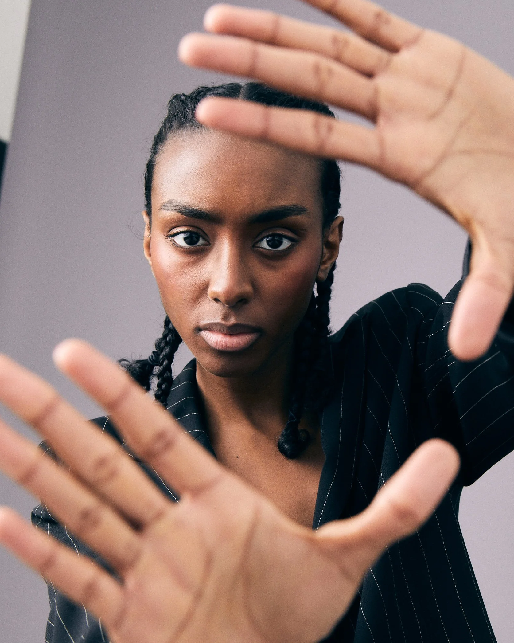 A woman with braided hair and a serious expression, wearing a black pinstripe shirt, is holding up her hands in front of the camera, partially obscuring her face.