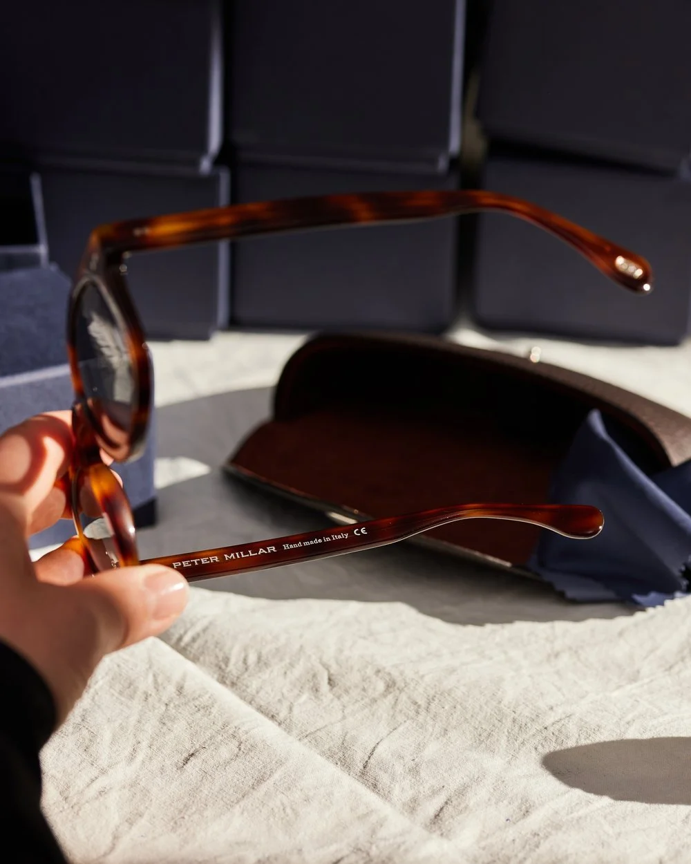 Close-up of brown tortoiseshell eyeglasses with the text "PETER MILLAR Handmade in Italy" on one arm, held in hand against a backdrop of dark boxes and an open case, with sunlight casting shadows.