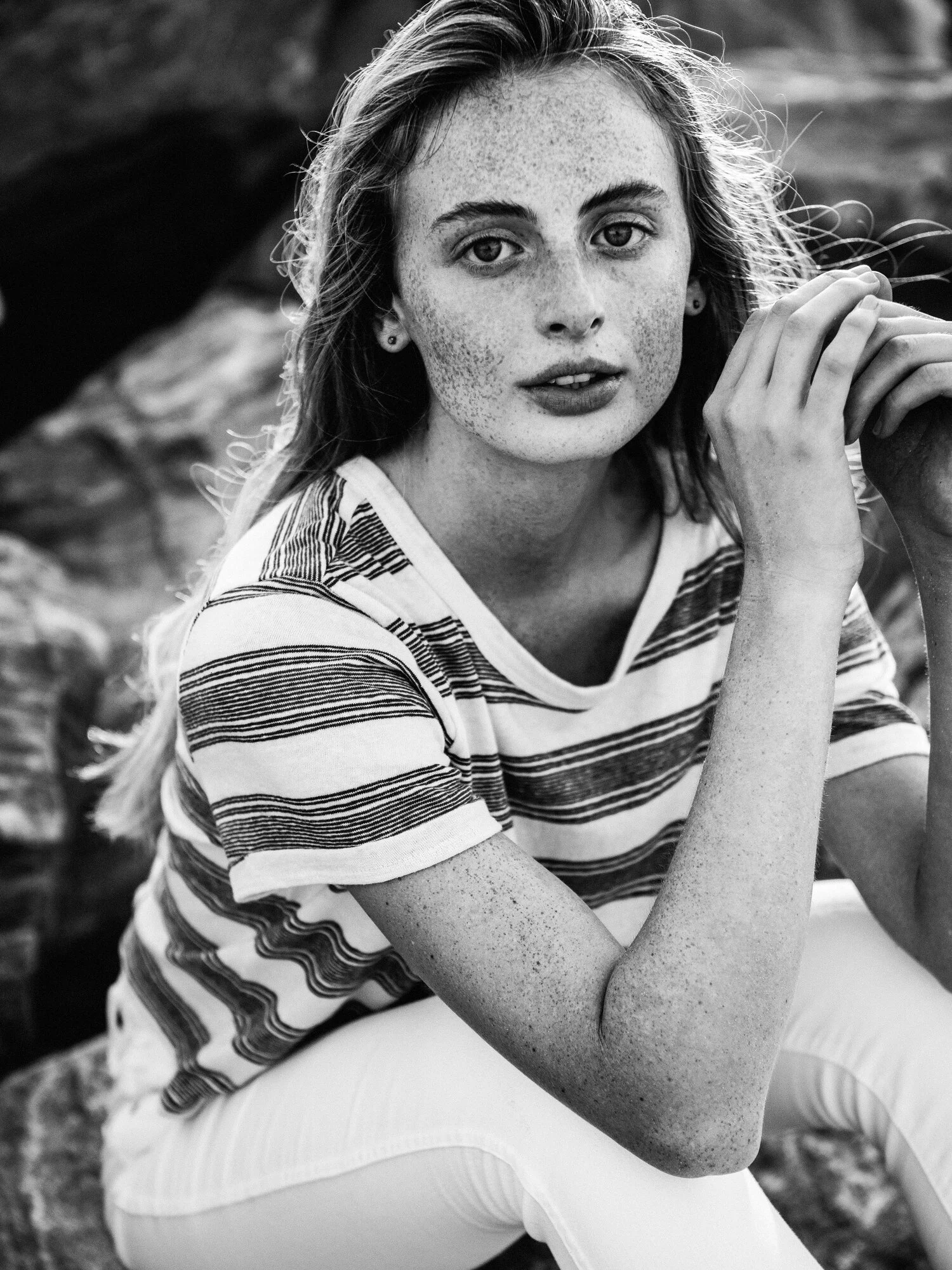 A young woman with curly hair and freckles, wearing a striped t-shirt and white pants, sitting outdoors near rocks, looking into the camera.