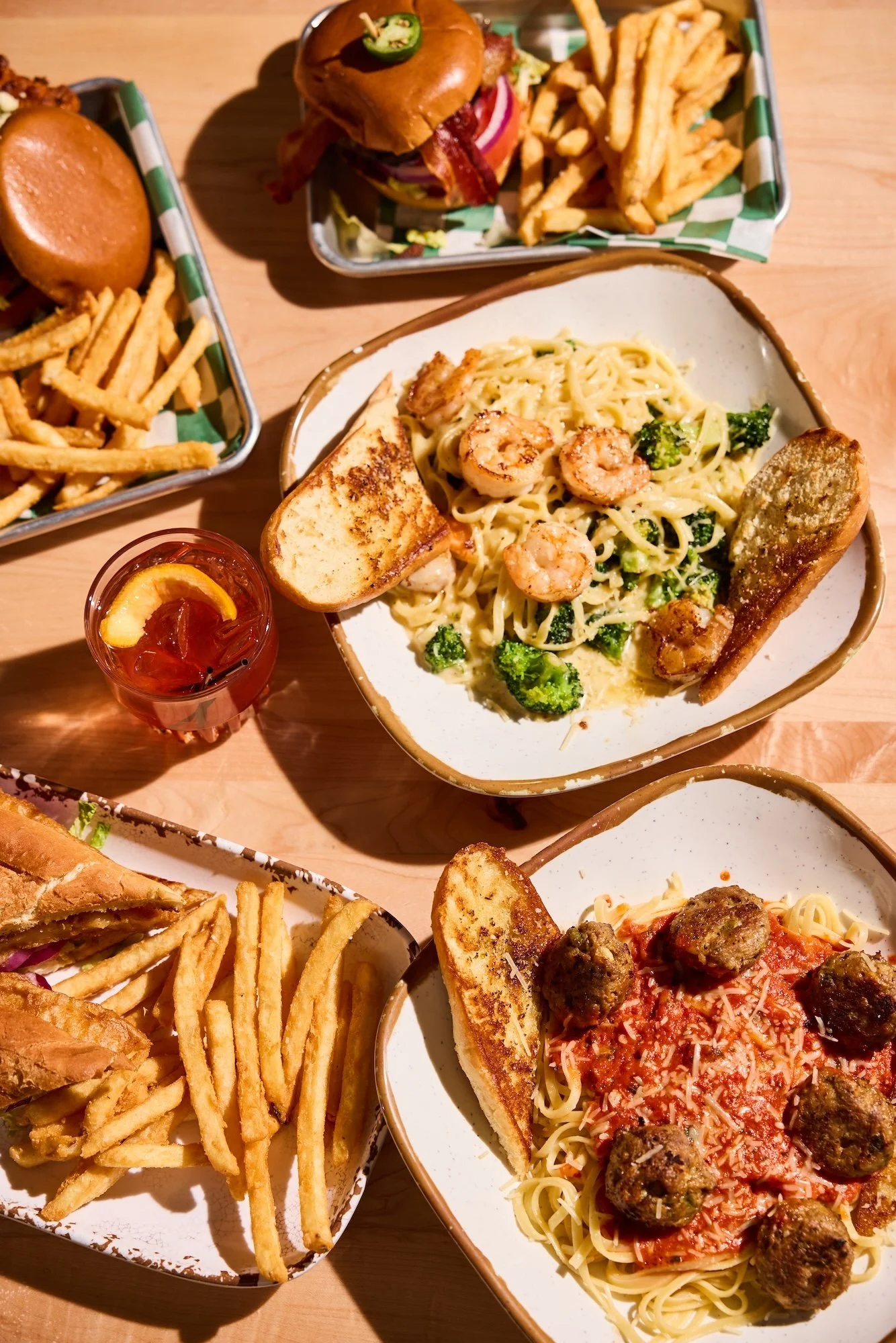 A table with two dishes of servings of pasta with shrimp and broccoli, topped with bread and garlic bread, along with two trays of French fries, and a glass of iced tea with lemon.