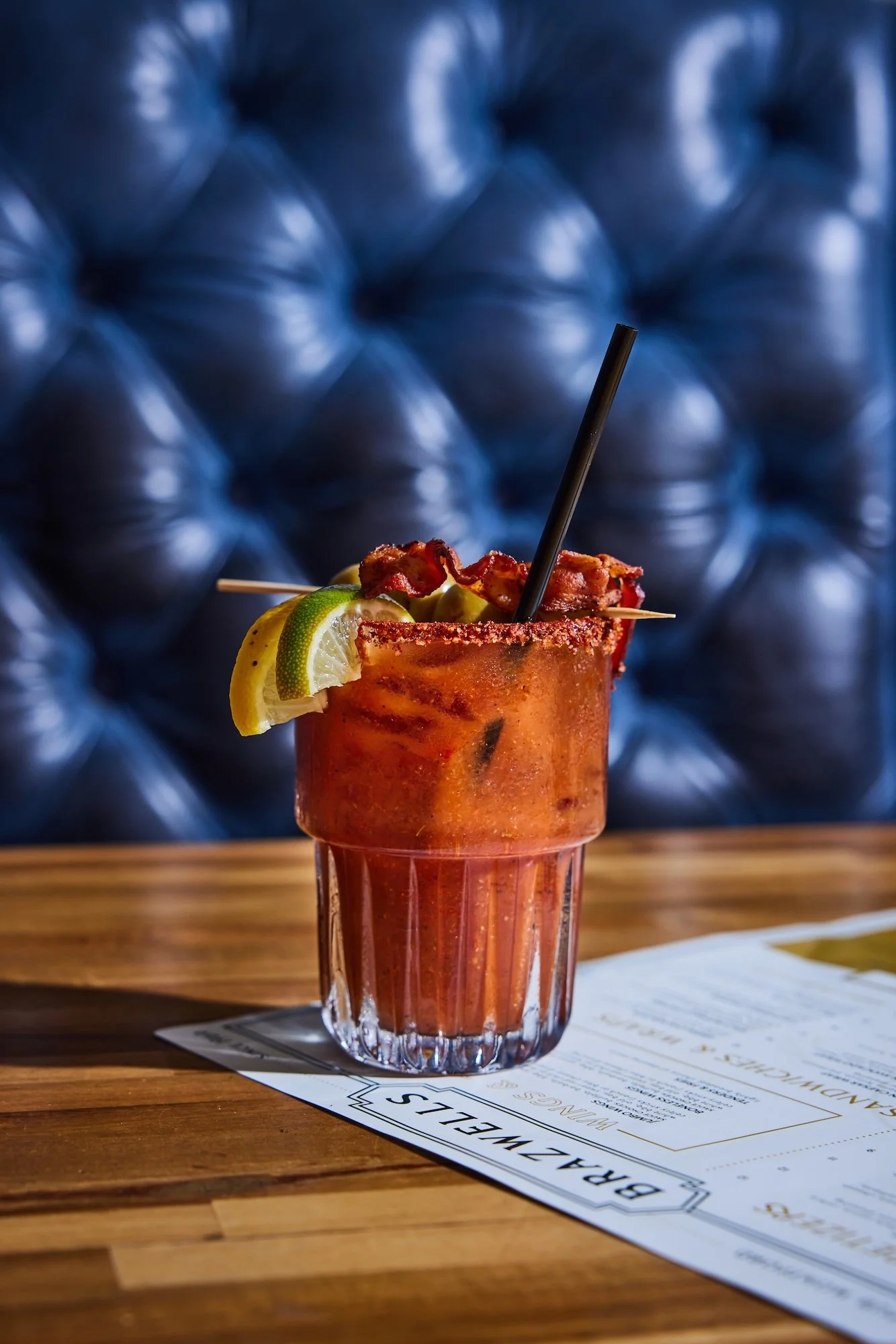 A Bloody Mary cocktail garnished with a lemon and lime wedge and a strip of bacon on a skewer, on a wooden table with a bar menu underneath, against a blue tufted background.