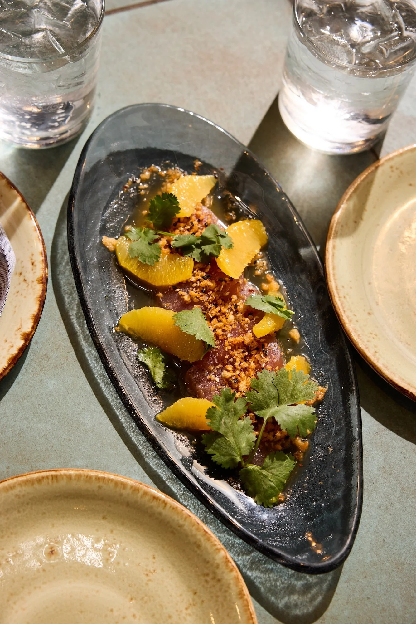 A black oval platter with thin slices of raw fish topped with orange slices, chopped nuts or breadcrumbs, and garnished with cilantro leaves, accompanied by two glasses of ice water and empty beige bowls.