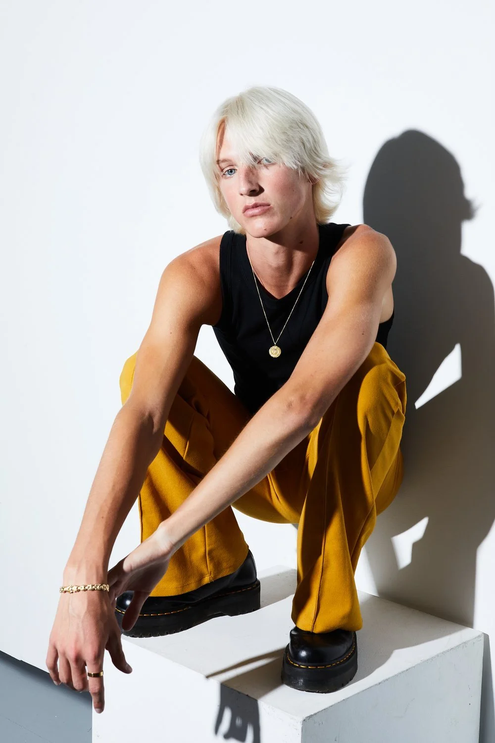 Person with platinum blonde hair squatting on a white platform wearing a black sleeveless top, yellow pants, and black shoes, casting a shadow on the white background.