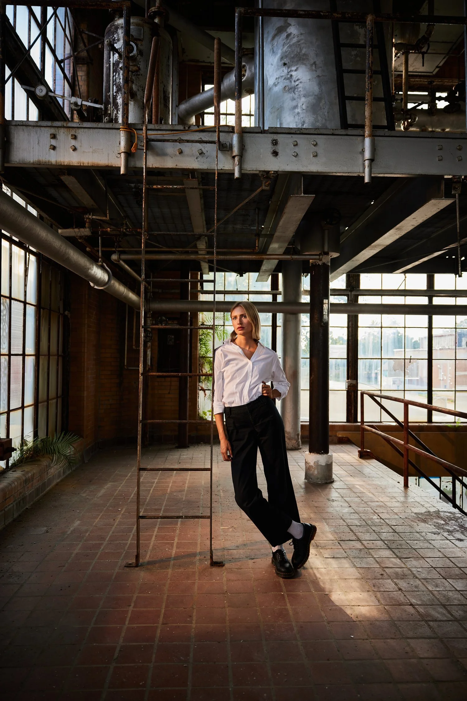 A young woman with blond hair wearing a white shirt, black trousers, and black shoes, standing in an industrial-style space with large windows, metal pipes, and exposed ductwork.