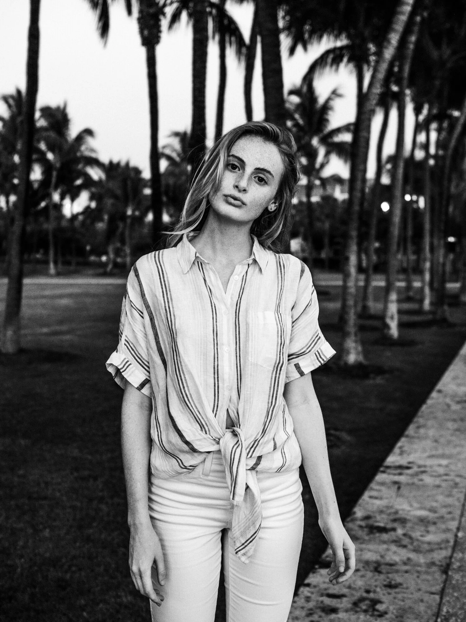 Black and white photo of a young woman with shoulder-length hair, wearing a striped button-up shirt tied at the waist and light-colored pants, standing outdoors near palm trees.