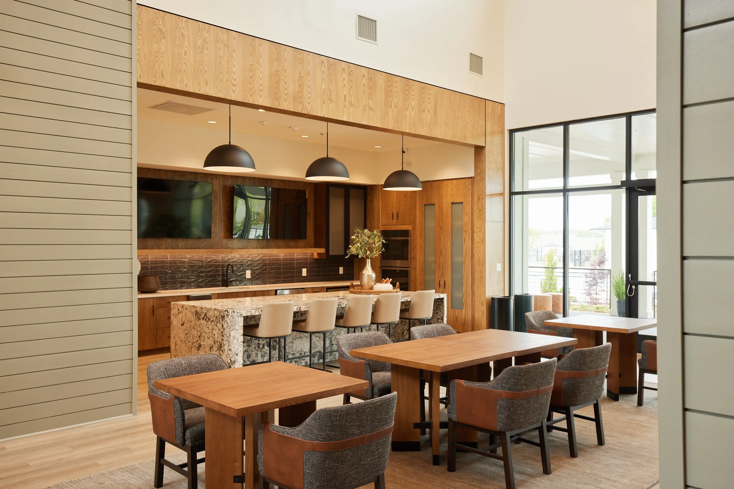 Modern kitchen and dining area with wooden tables and upholstered chairs, large windows, and a kitchen island with barstools and pendant lighting.