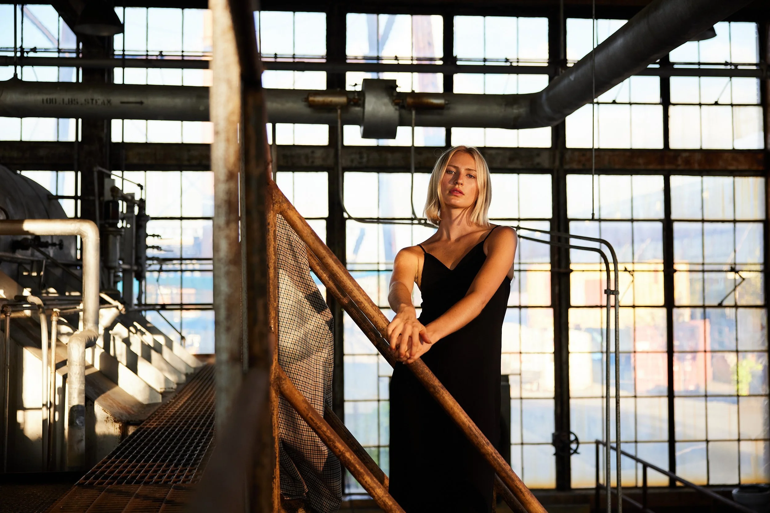 A woman with blonde hair in a black dress standing on an industrial metal staircase inside a factory with large windows and pipes.