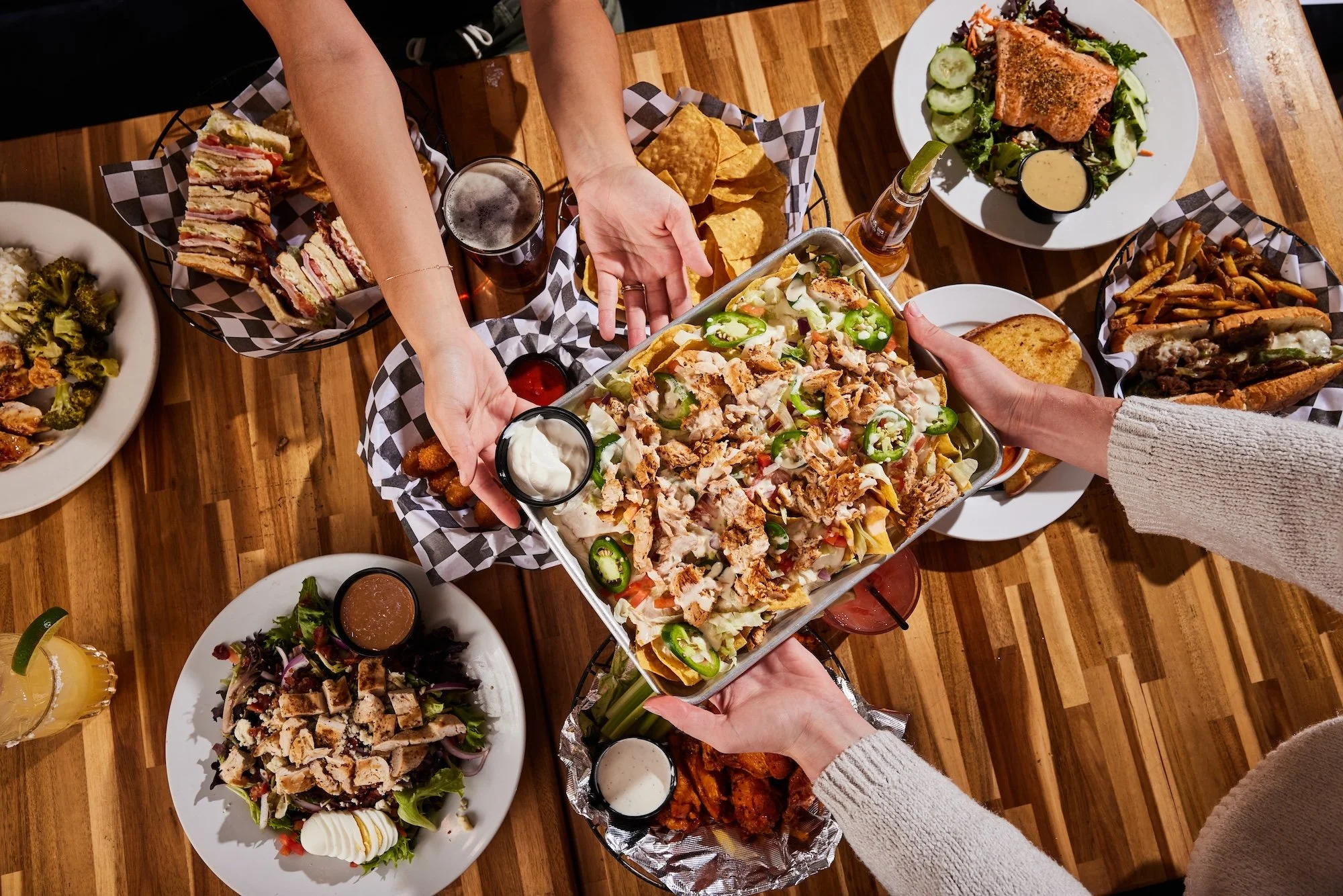 People passing around a variety of dishes on a wooden table, including salads, sandwiches, nachos, wings, ribs, and a platter with shredded chicken and jalapenos.