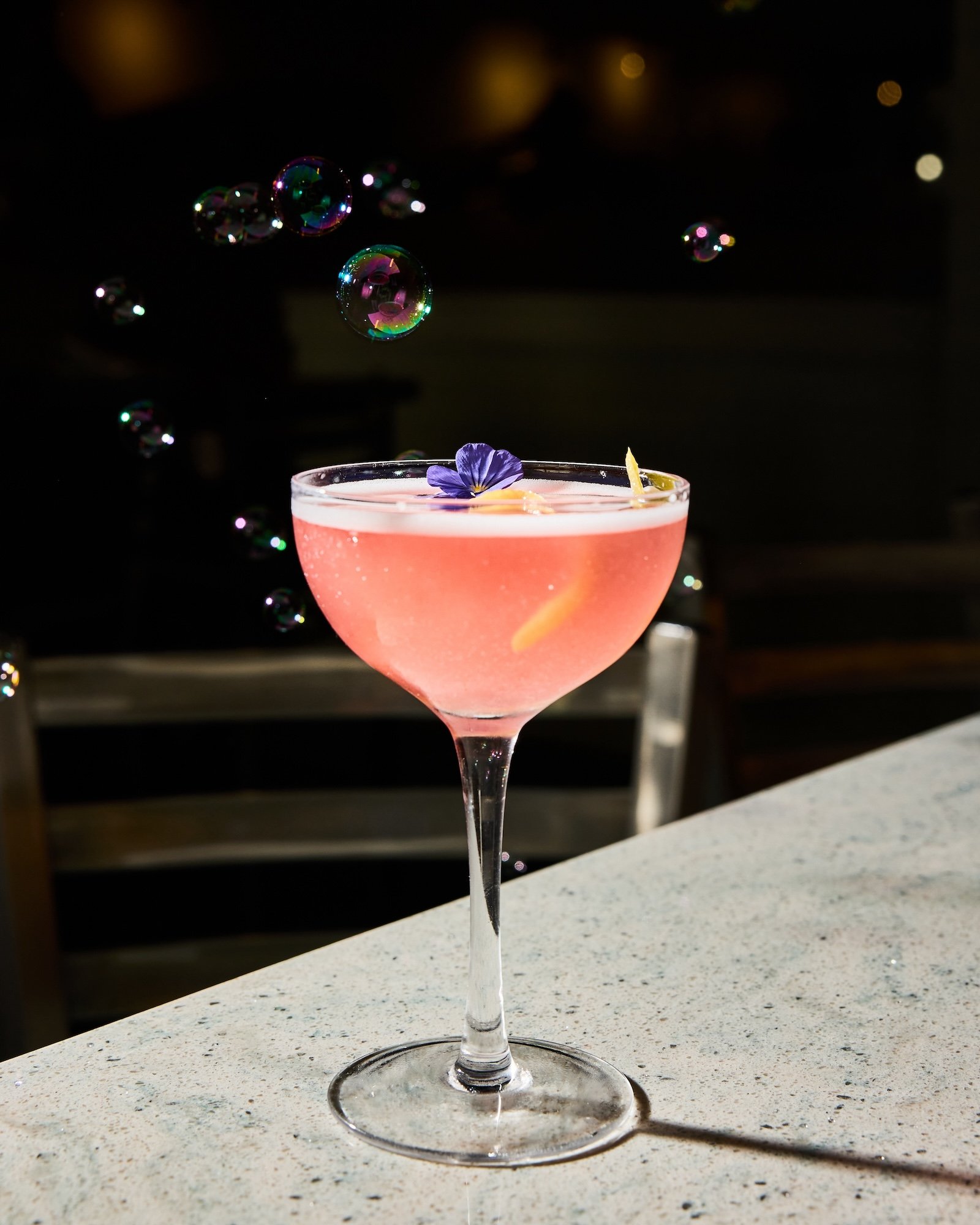 A pink cocktail garnished with an edible flower in a coupe glass on a white speckled surface, with soap bubbles floating above it and a dark, blurred background.