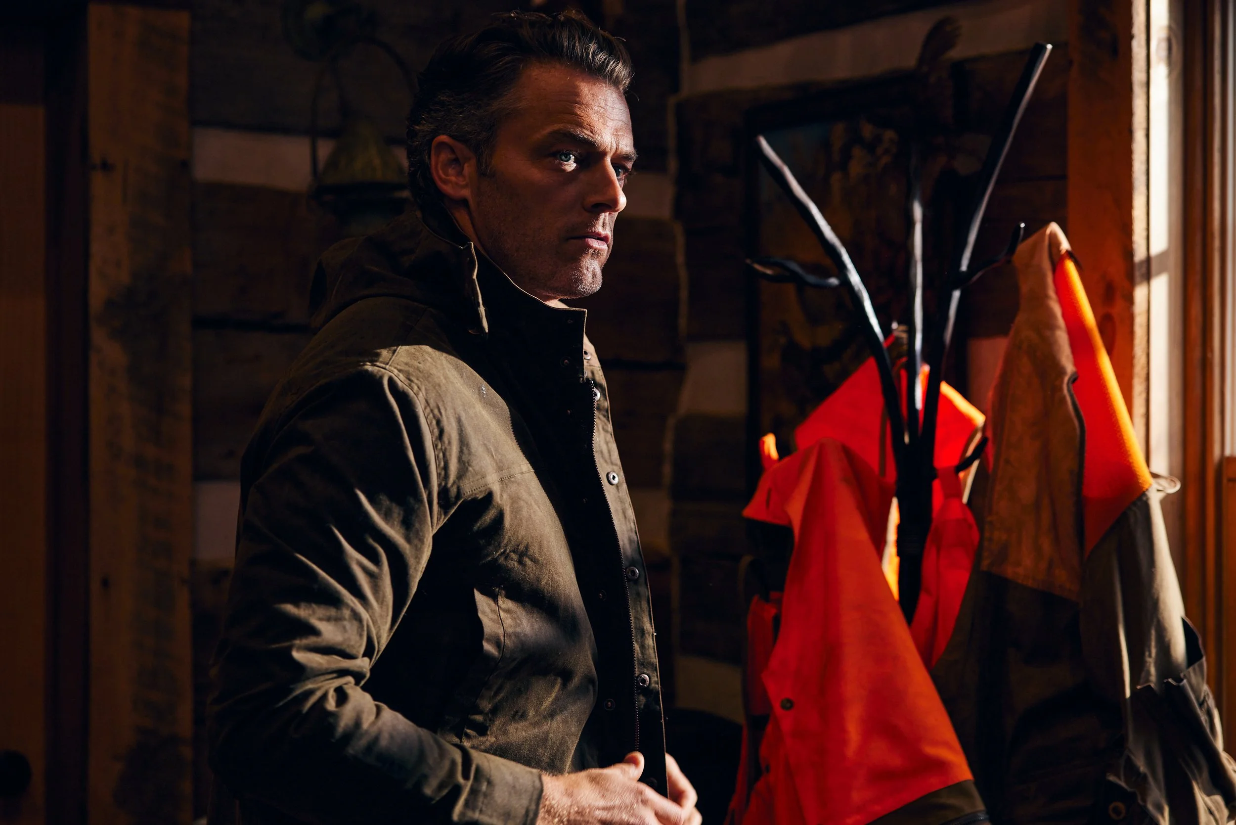 A man with dark hair and a serious expression standing indoors near a wooden wall, wearing a brown jacket, with outdoor gear hanging on a coat rack beside him.