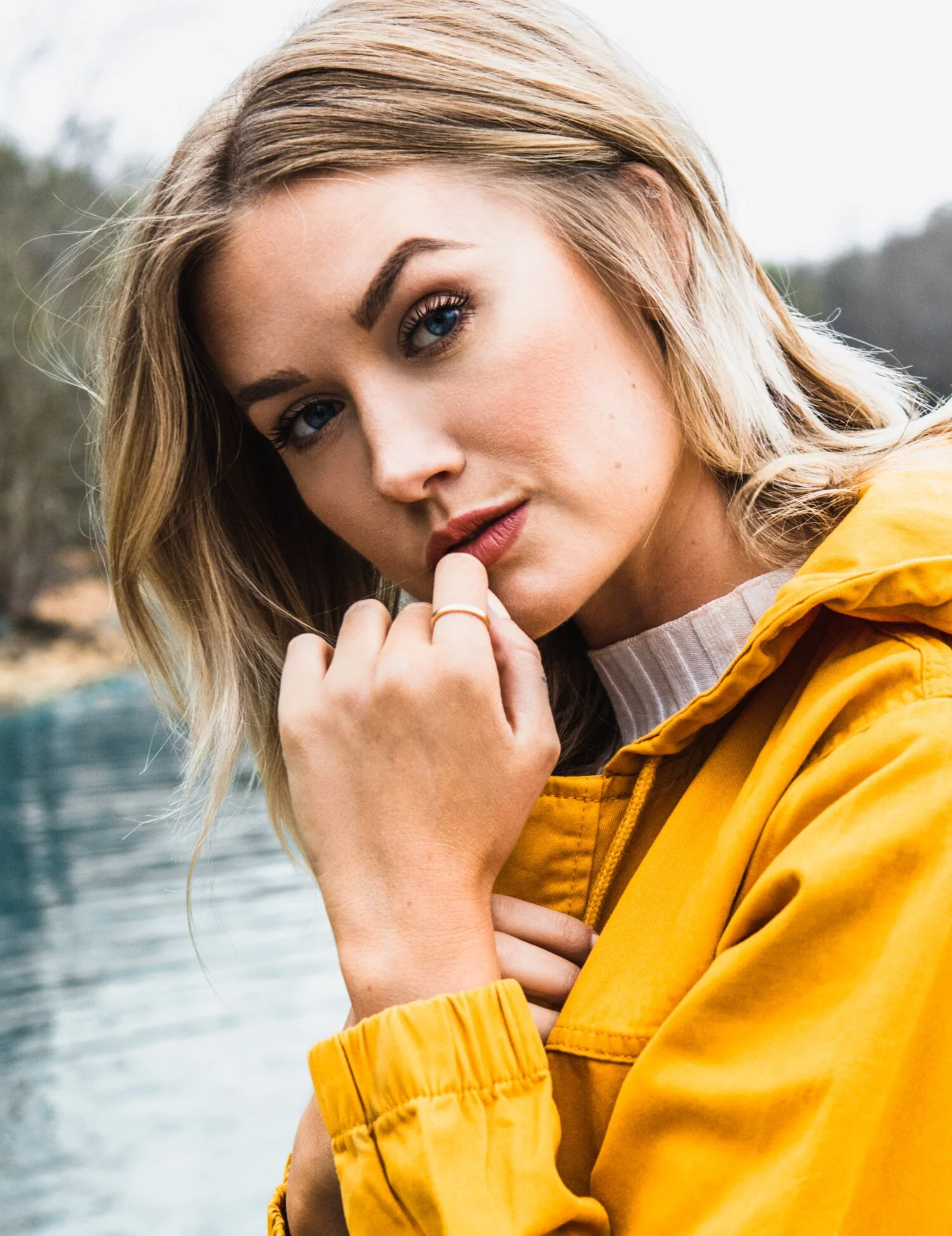 A young woman with blonde hair and blue eyes, wearing a yellow jacket, poses outdoors near water with trees in the background, touching her lips with her fingers.