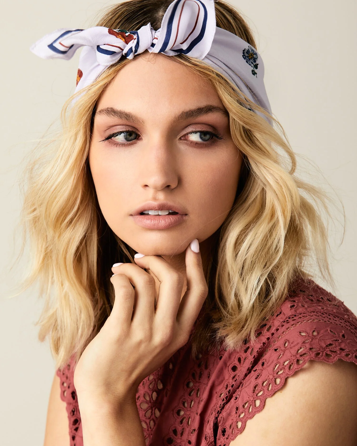 Close-up portrait of a woman with blonde wavy hair, wearing a white headband with a blue and red pattern, and a pink lace top. She is looking to the side with her hand gently touching her chin.