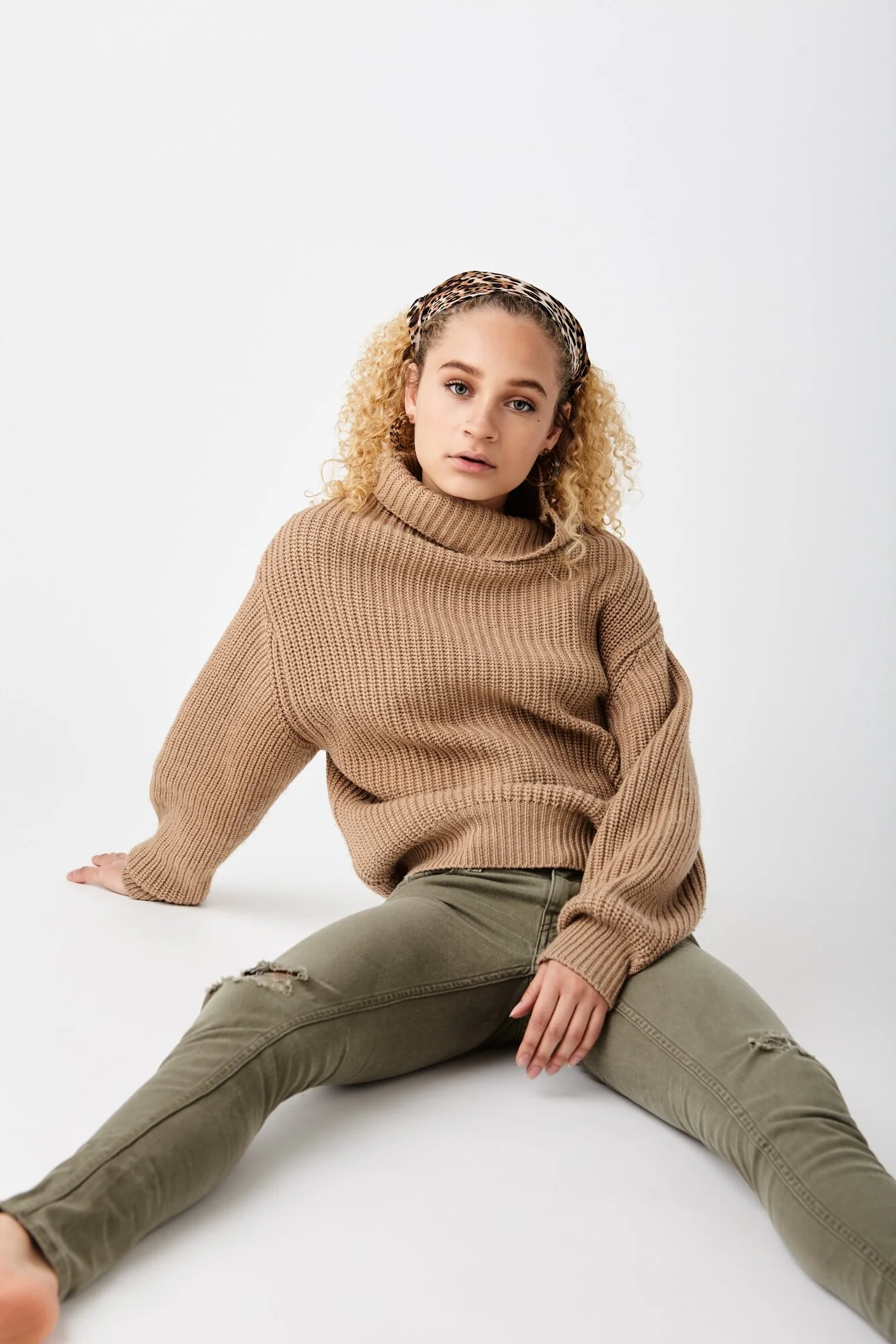 A young woman with curly blonde hair wearing a tan sweater, olive green ripped jeans, and a leopard print headband, sitting on the floor against a plain white background.