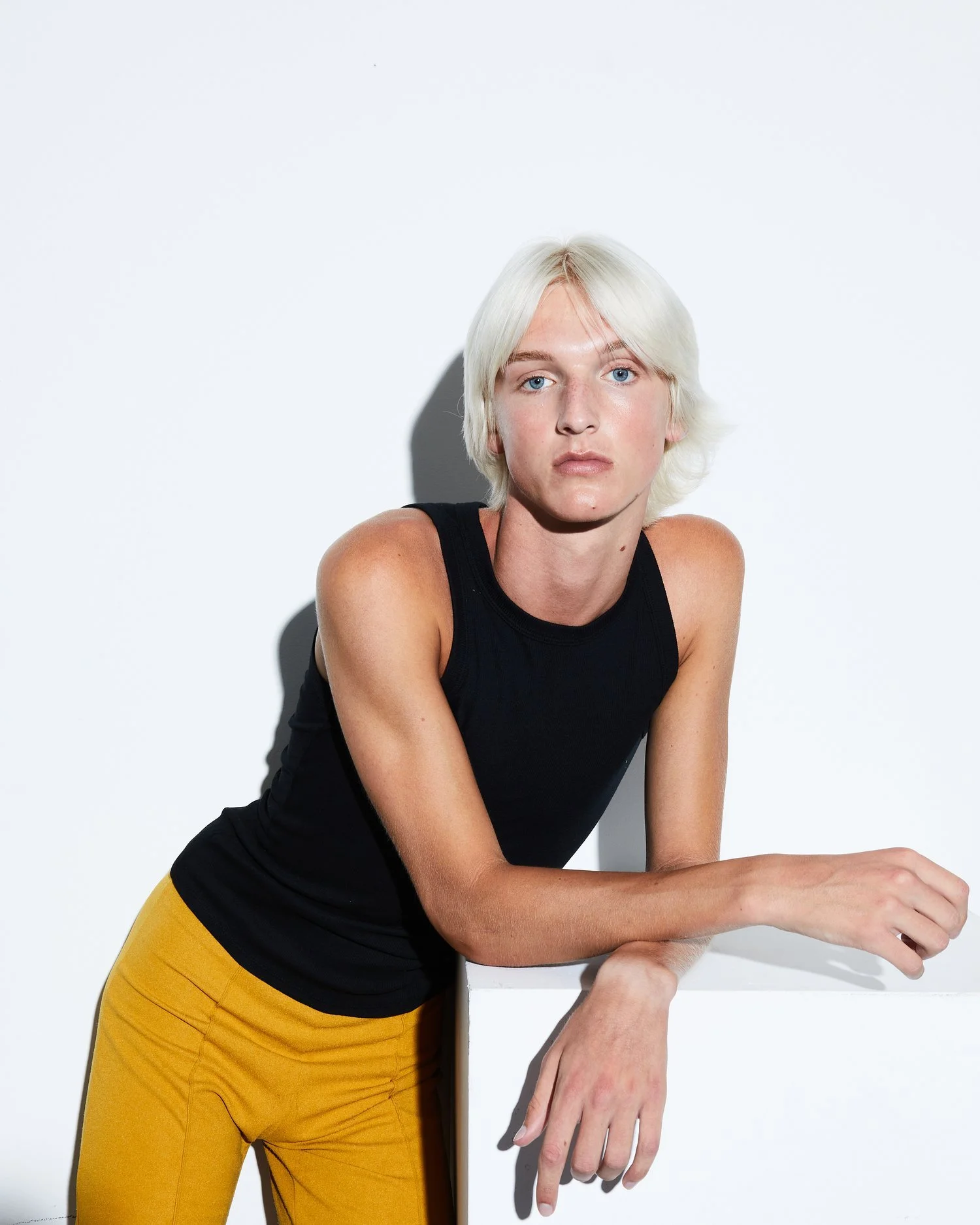 Young person with platinum blonde hair, wearing a black sleeveless top and yellow pants, leaning on a white surface against a white background.