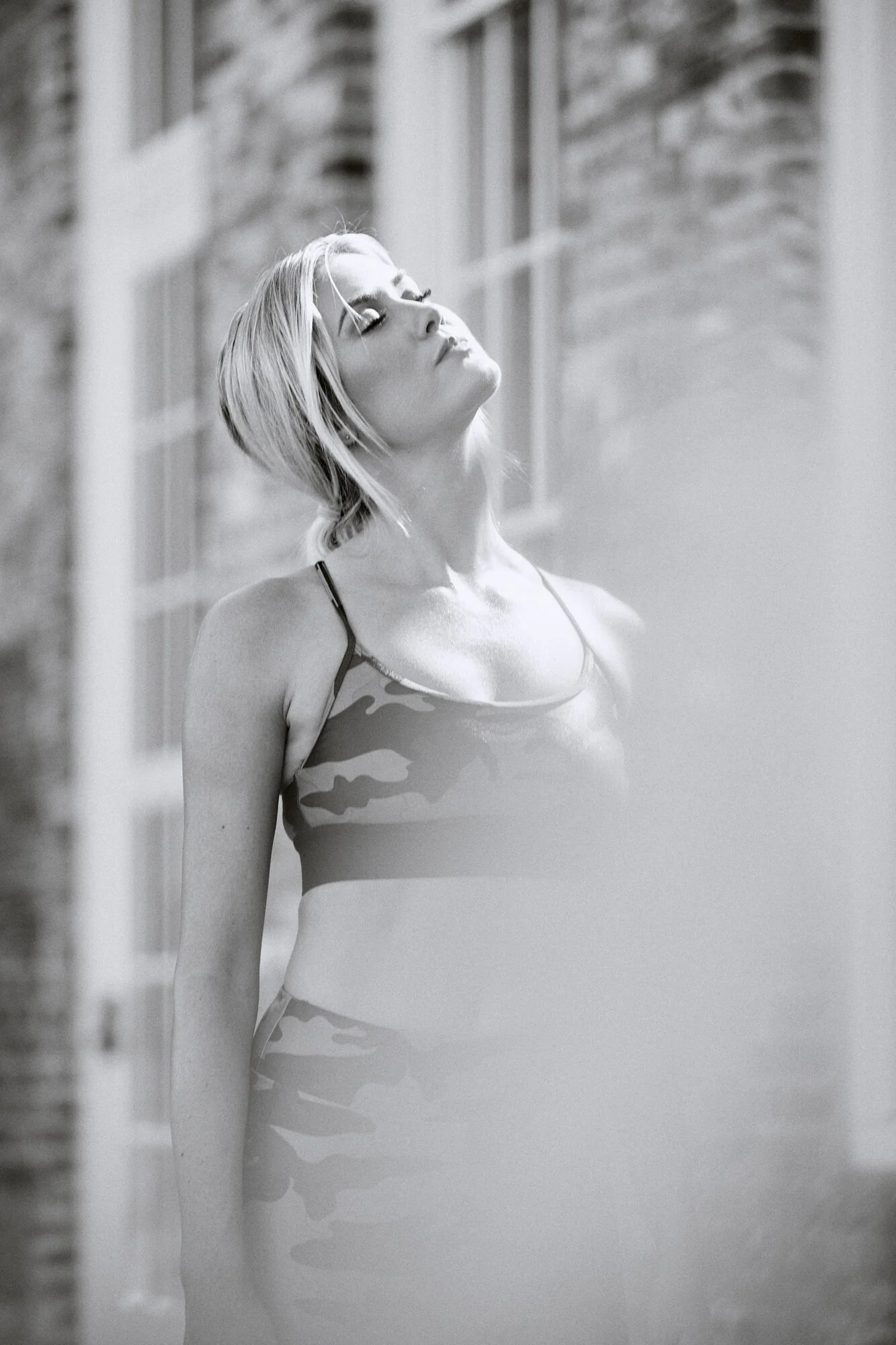 A woman with short blonde hair in a sports bra and leggings, standing outdoors with closed eyes and a relaxed expression, near a brick building in black and white.