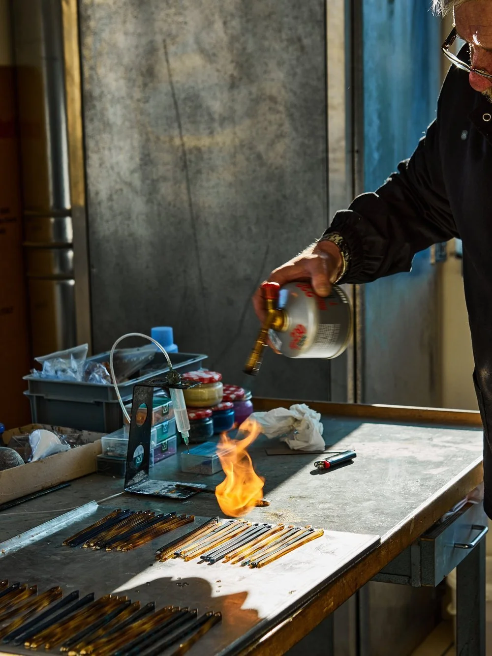 A person working with glass rods, using a flame and chemicals, in a glassmaking workshop.