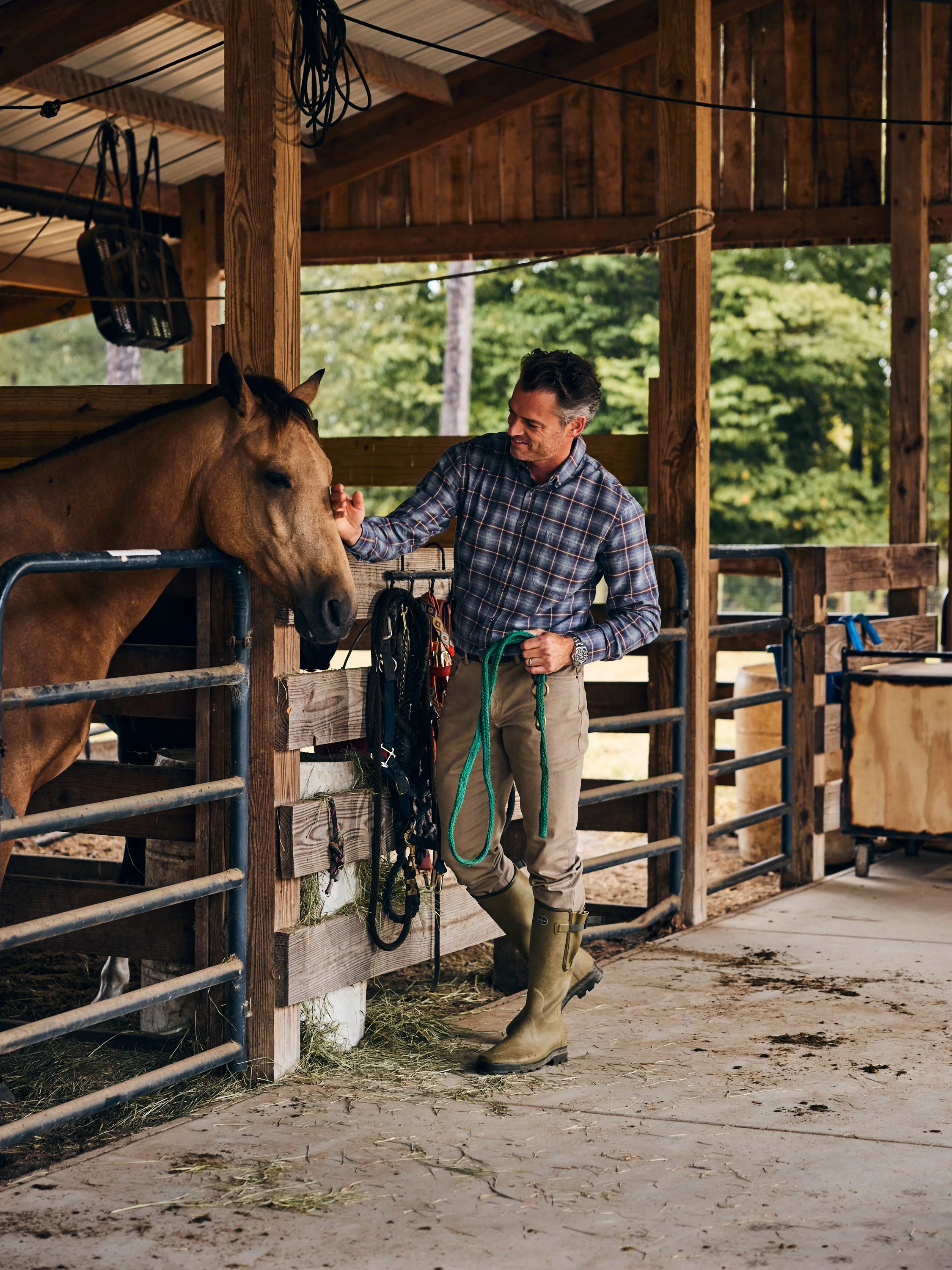 A man in plaid shirt and rubber boots petting a brown horse inside a wooden stable.
