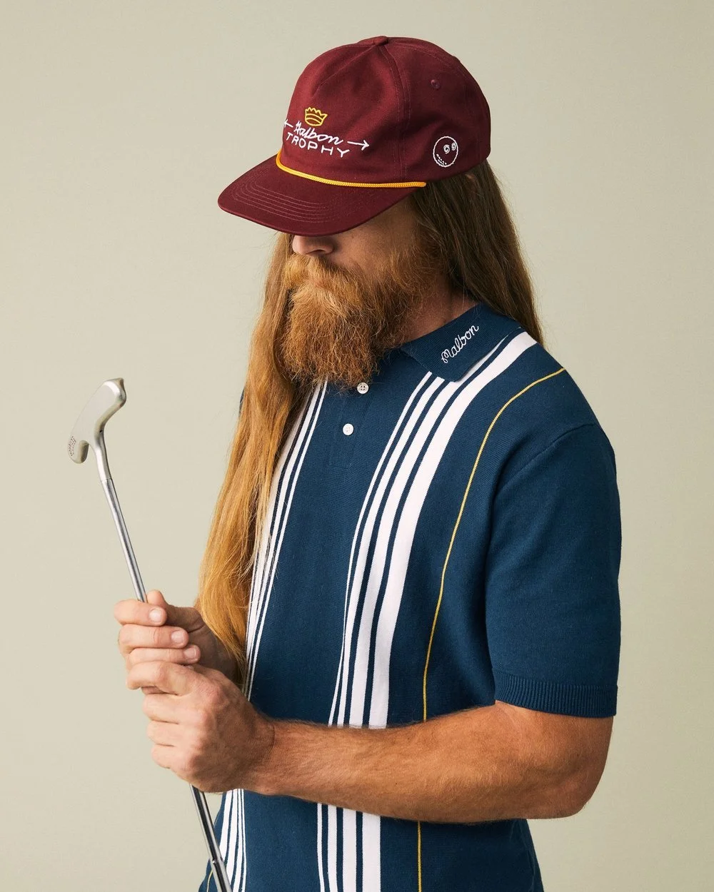A man with long hair and a beard wearing a navy blue golf shirt and a red cap looking at a golf club.