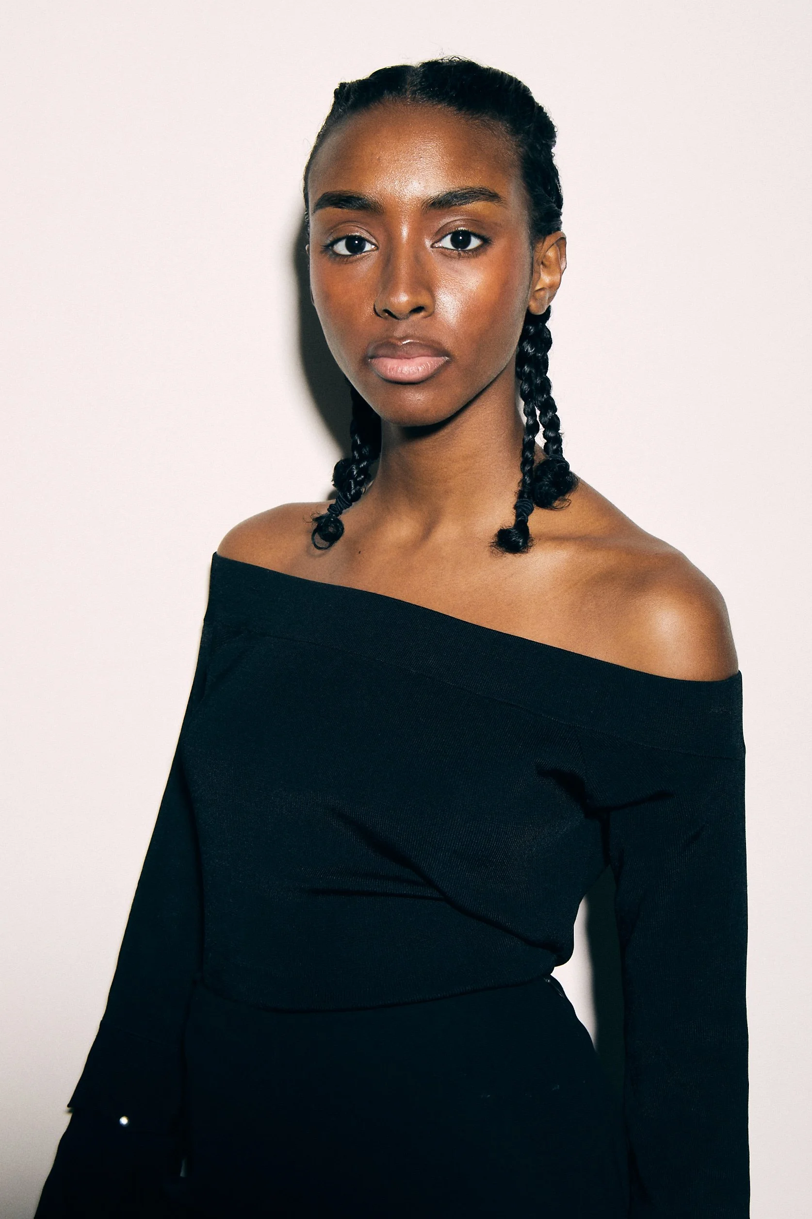 Portrait of a woman with braided hair wearing an off-the-shoulder black top against a plain background.