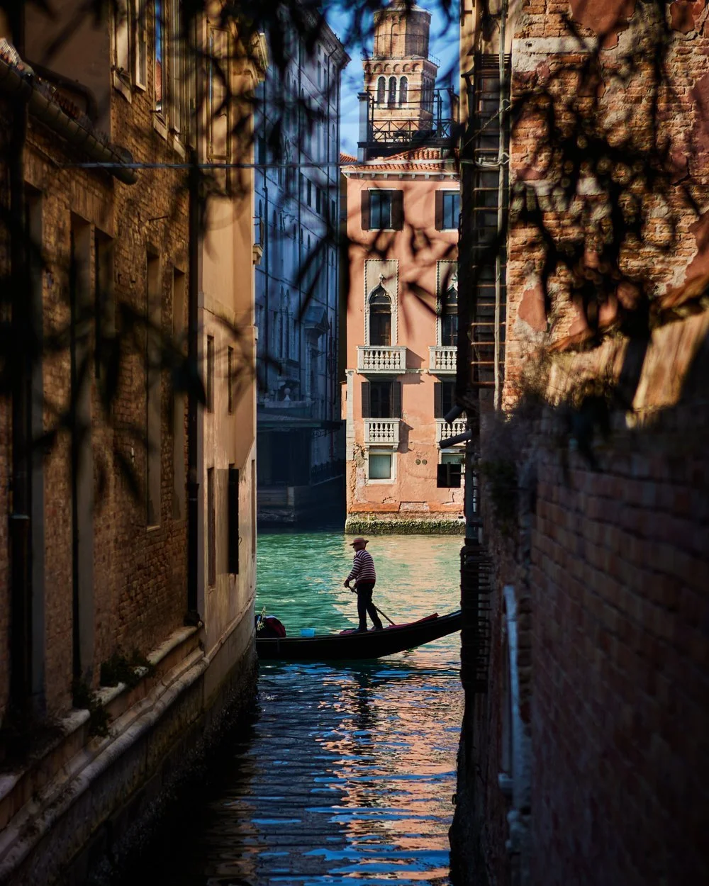 A gondolier in striped shirt standing on a gondola navigating through a narrow canal between old brick buildings in Venice, Italy, with a brick tower in the background.