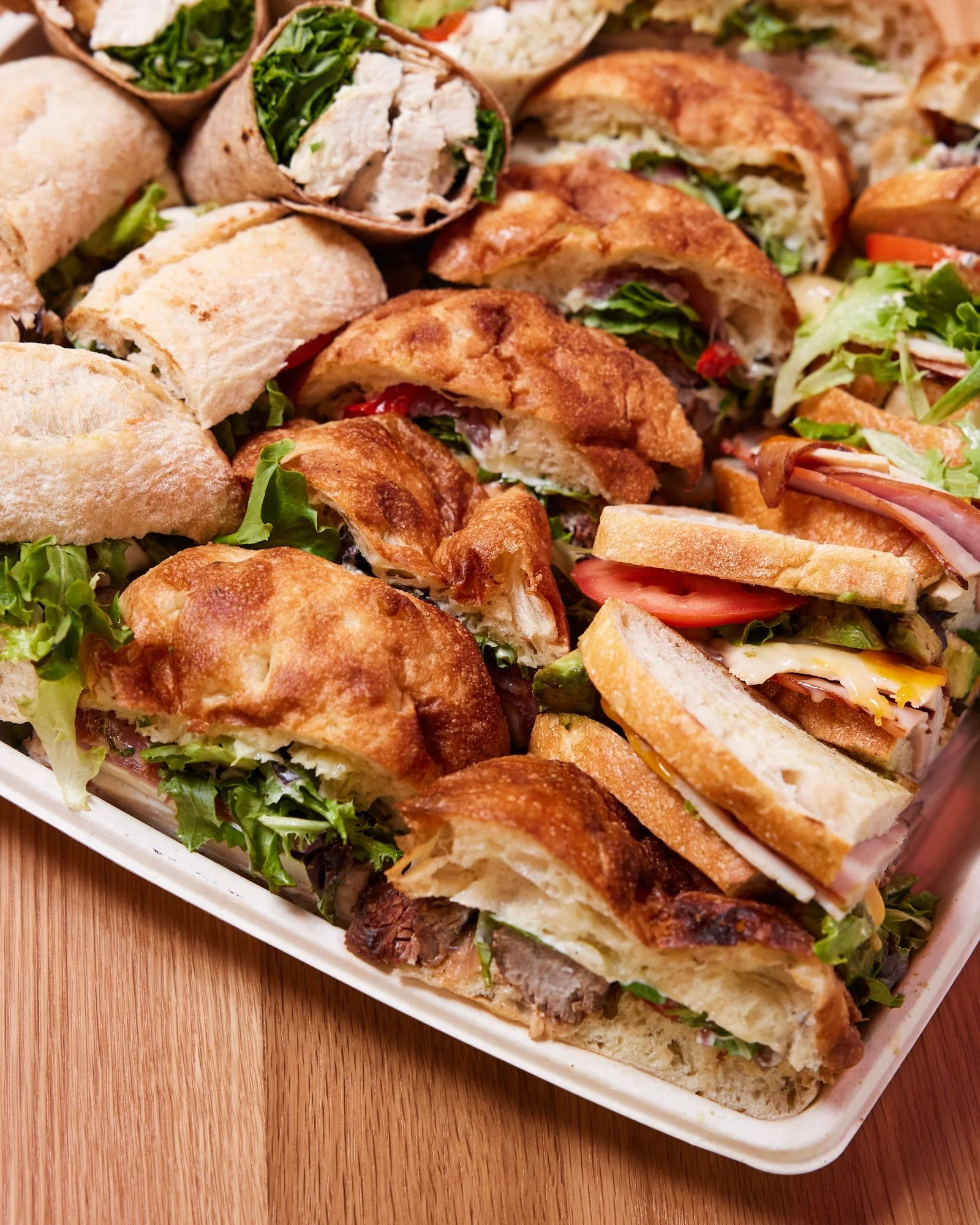 A tray of assorted sandwiches including wraps, sub sandwiches, and croissant sandwiches filled with vegetables, meats, and cheese, served on a white platter on a wooden surface.