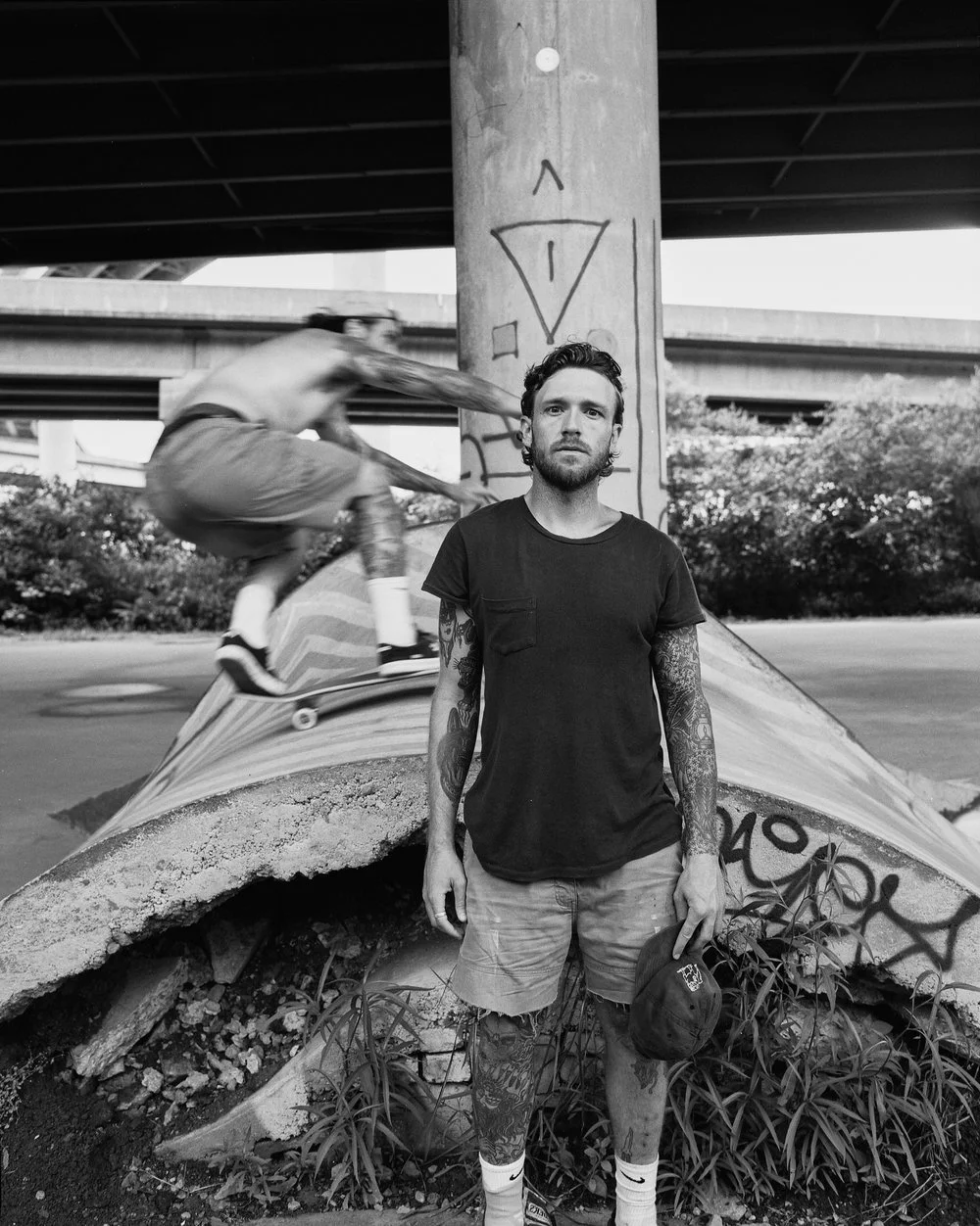 A man with tattoos wearing a black t-shirt and shorts standing under a highway overpass, holding a baseball cap. A skateboarder is jumping over a broken concrete structure behind him. The scene is urban with graffiti on the support pillar and overpas