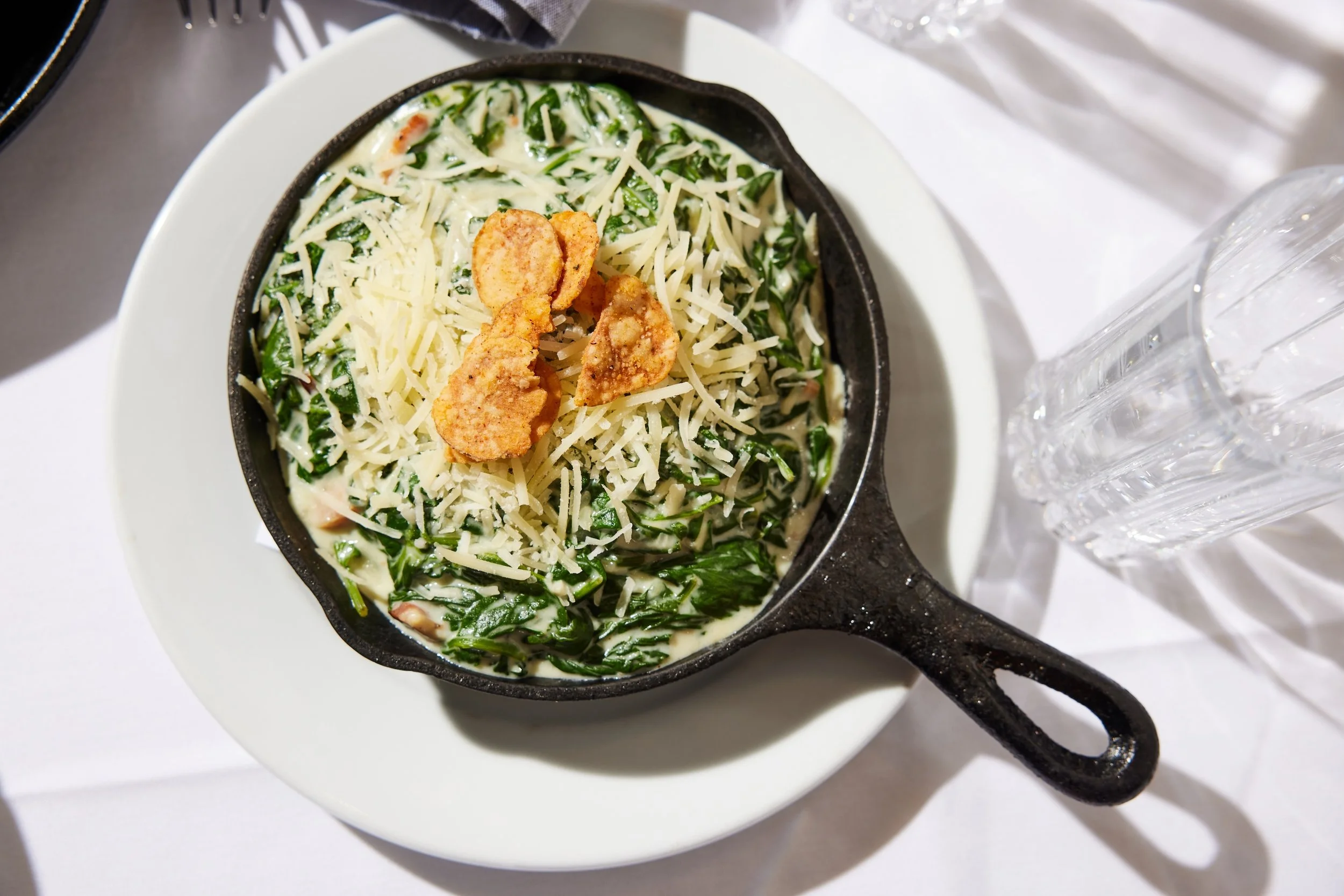Creamy spinach dip topped with shredded cheese and fried garlic chips in a small cast iron skillet over a white plate.