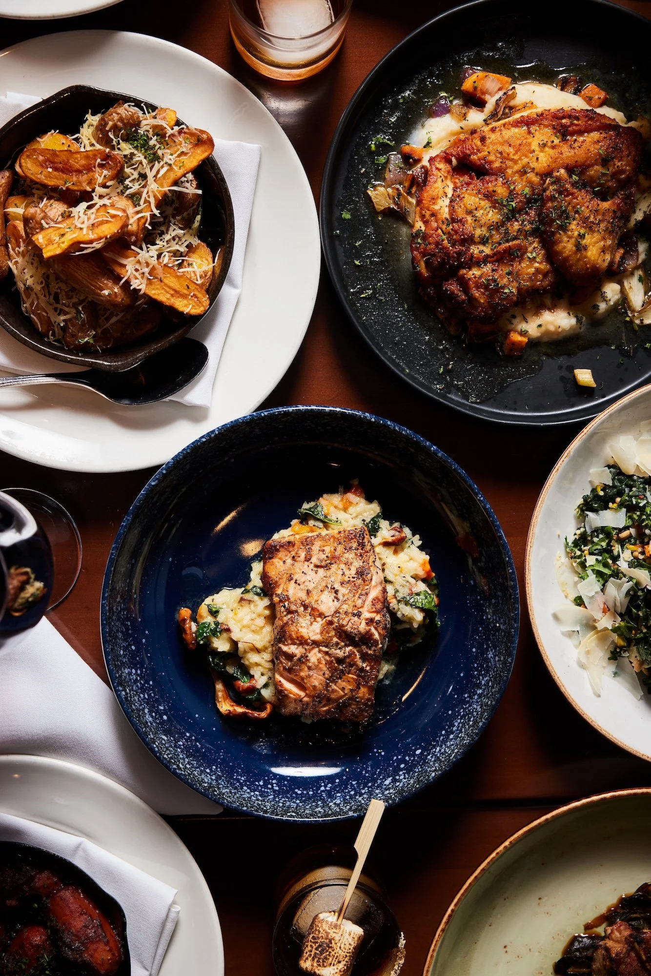 A variety of plated gourmet dishes including roasted chicken, mashed potatoes with a pork chop, roasted potatoes with herbs, and a salad, along with glasses of beer and wine on a wooden table.