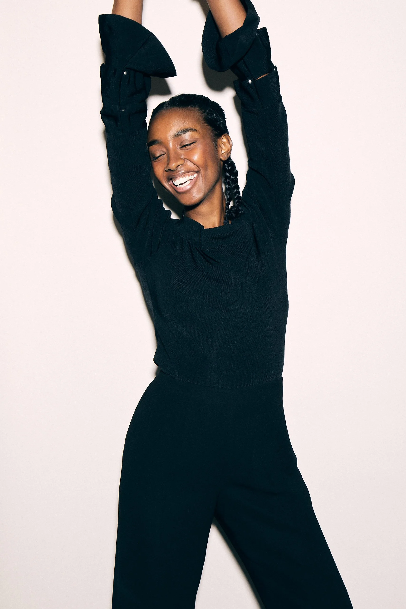 A woman with dark hair styled in braids, wearing a black long-sleeve top and black pants, smiling with eyes closed and arms raised, standing against a plain white background.