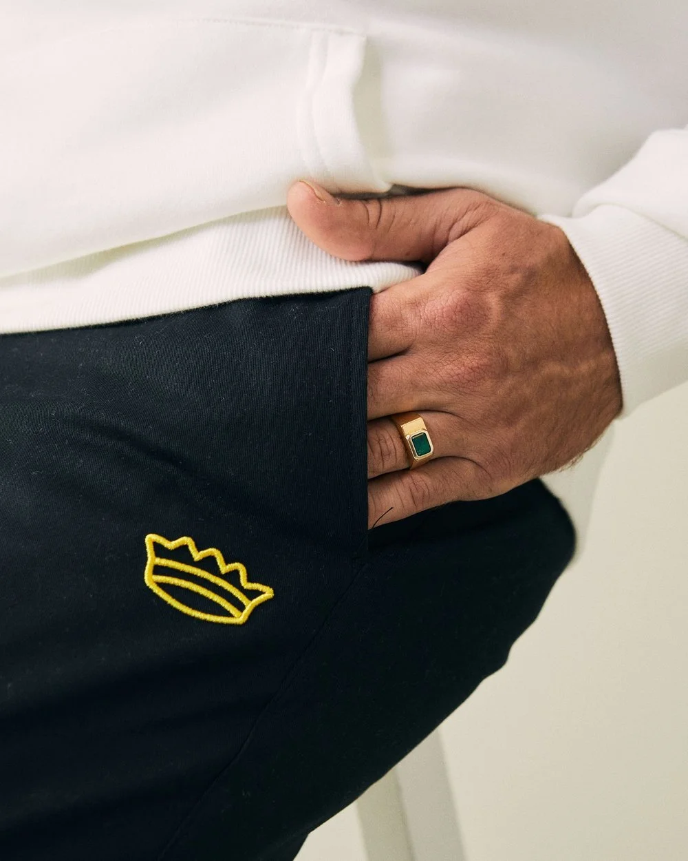 Close-up of a person's hand resting on their hip, wearing a gold ring with a green stone, with a white sweatshirt and black pants featuring a yellow embroidered logo.