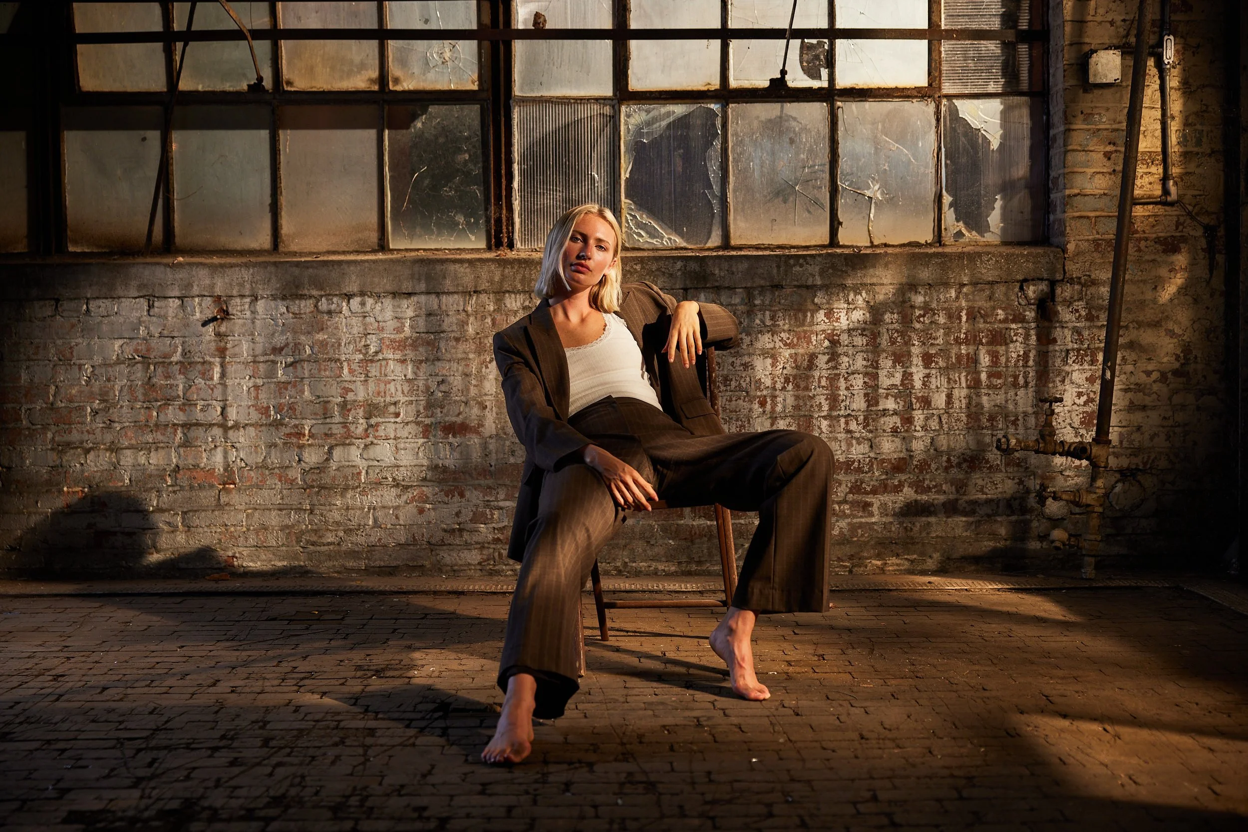 A woman in business attire sitting barefoot in an industrial-style room with a brick wall and broken windows.