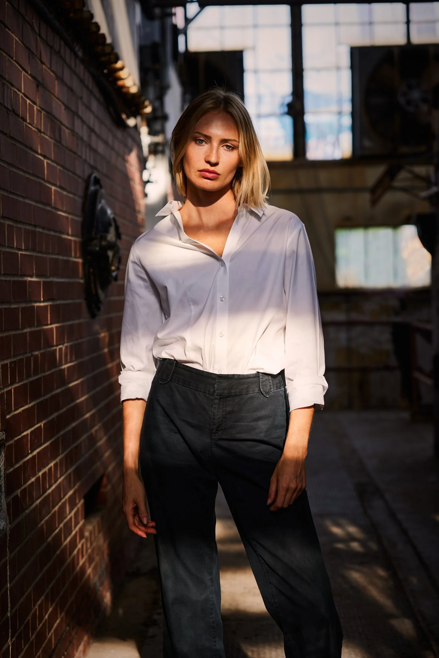A woman with blonde hair standing in an industrial-style space with brick walls and large windows, illuminated by natural light.