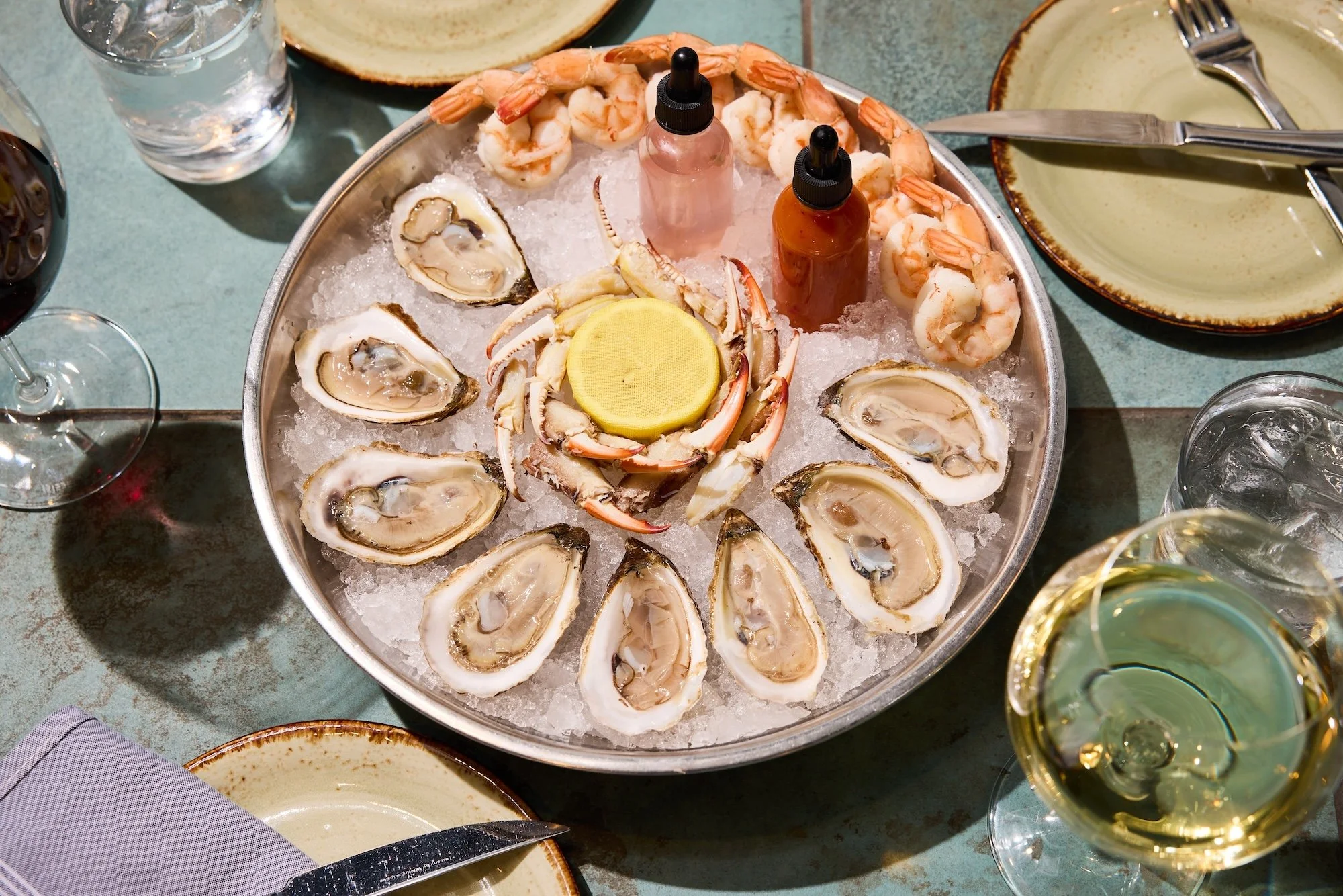 A seafood platter with oysters on the half shell, cooked shrimp, and crab claws on ice, garnished with lemon slices, accompanied by two bottles of hot sauce.