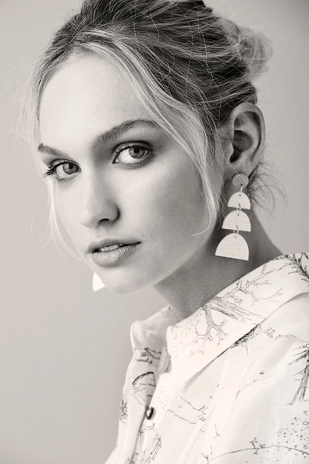 Black and white portrait of a young woman with light eyes and long hair styled in a loose updo, wearing large geometric earrings and a jacket with a map print.