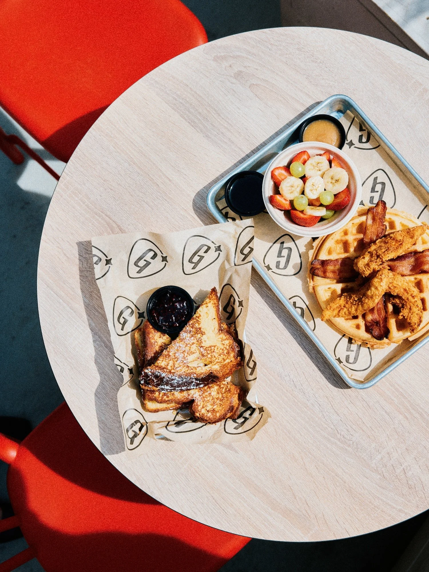 A breakfast meal on a round table includes French toast with a side of berry jam, a waffle topped with fried chicken and bacon, and a small bowl of fresh fruit with strawberries, bananas, and grapes, accompanied by two small containers of syrup.