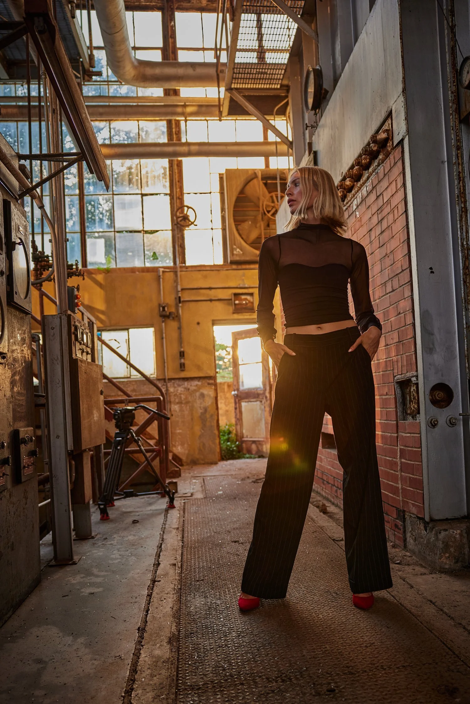 A woman with blonde hair wearing a black sheer top, pinstripe wide-leg pants, and red heels stands in an industrial setting with sunlight streaming through large windows.