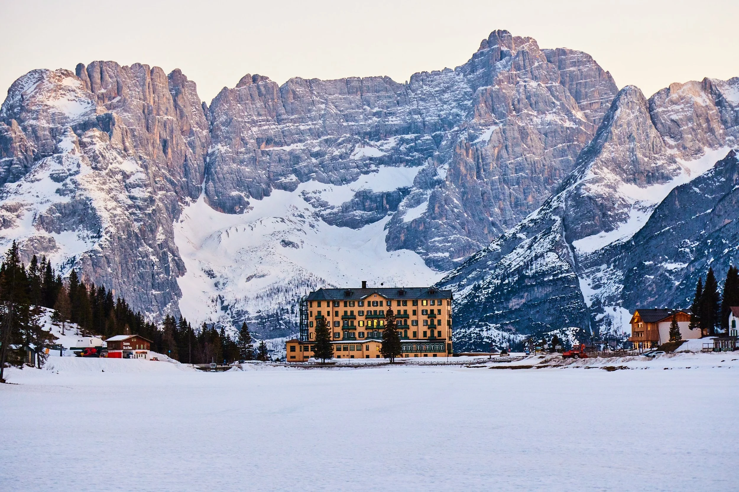 A large building with multiple floors is situated at the edge of a frozen lake with snow-covered mountains in the background.