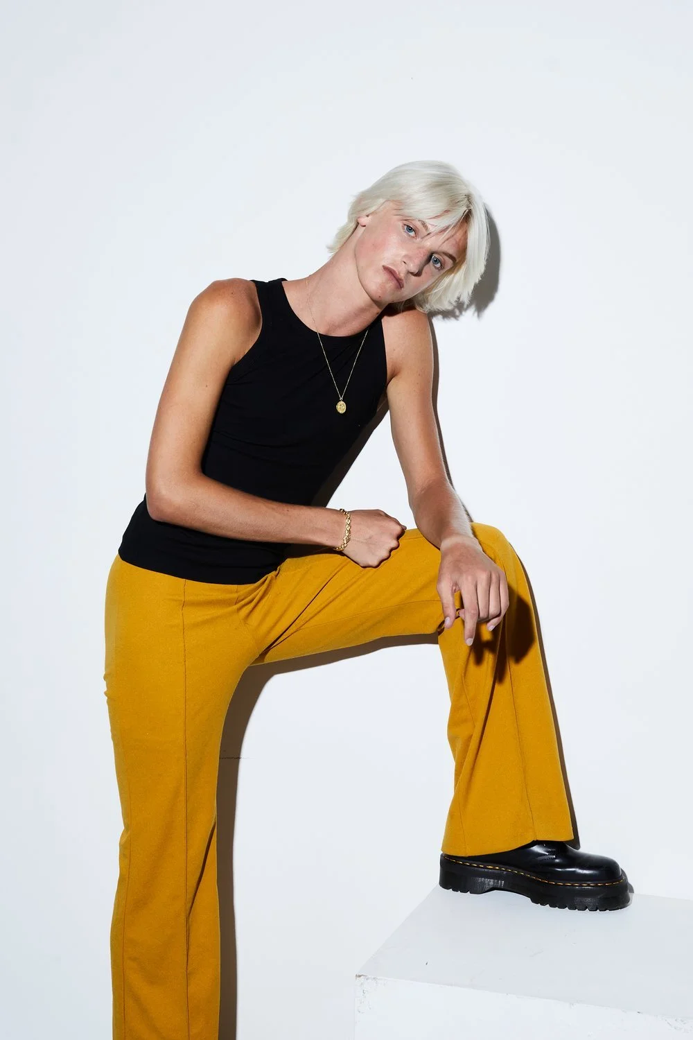 A person with short blonde hair wearing a black sleeveless top, mustard yellow pants, and black shoes, posing with one foot on a white platform against a white background.