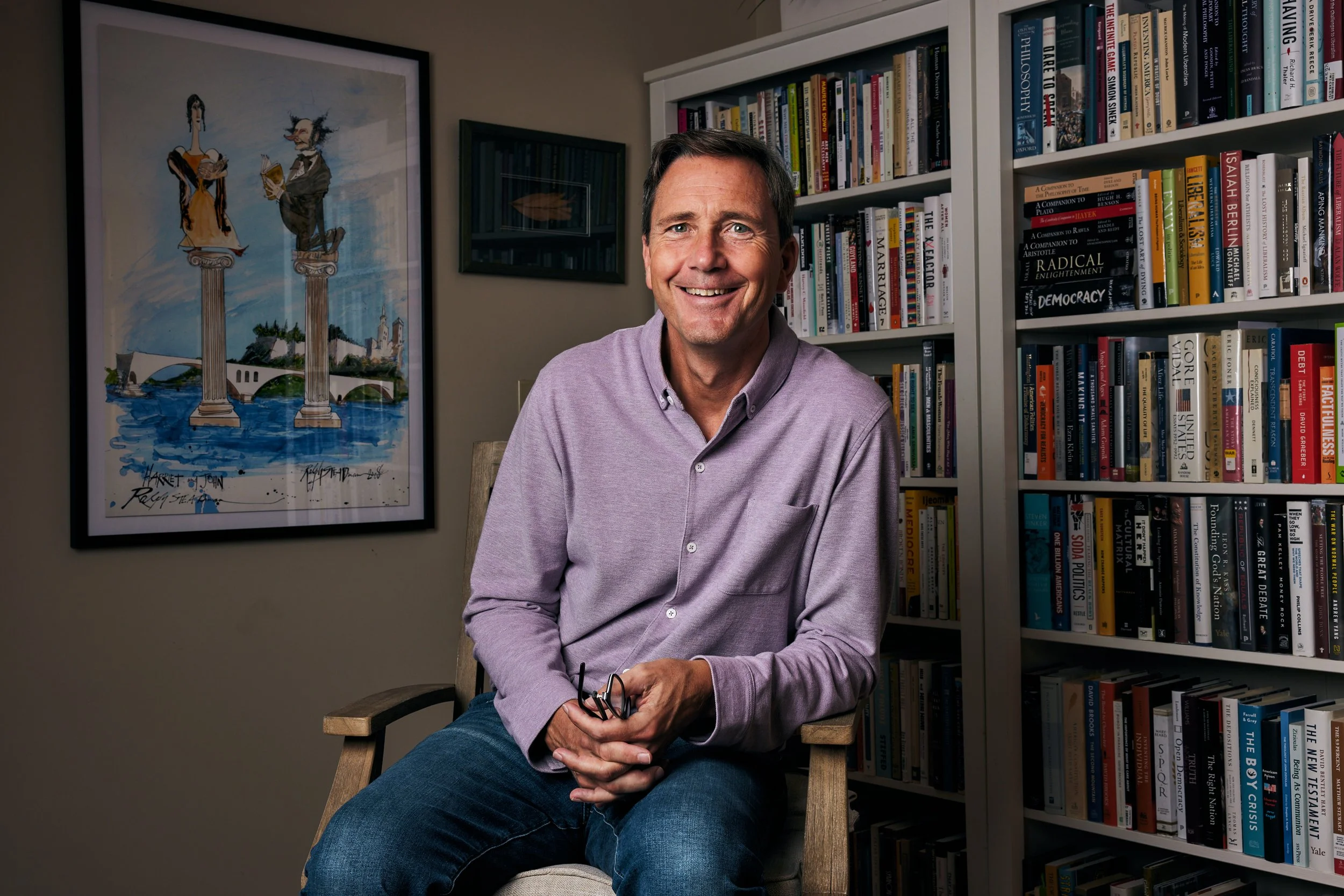 A smiling man sitting on a chair holding glasses in his hands, in a room with a large bookshelf filled with books and a framed cartoon art on the wall.