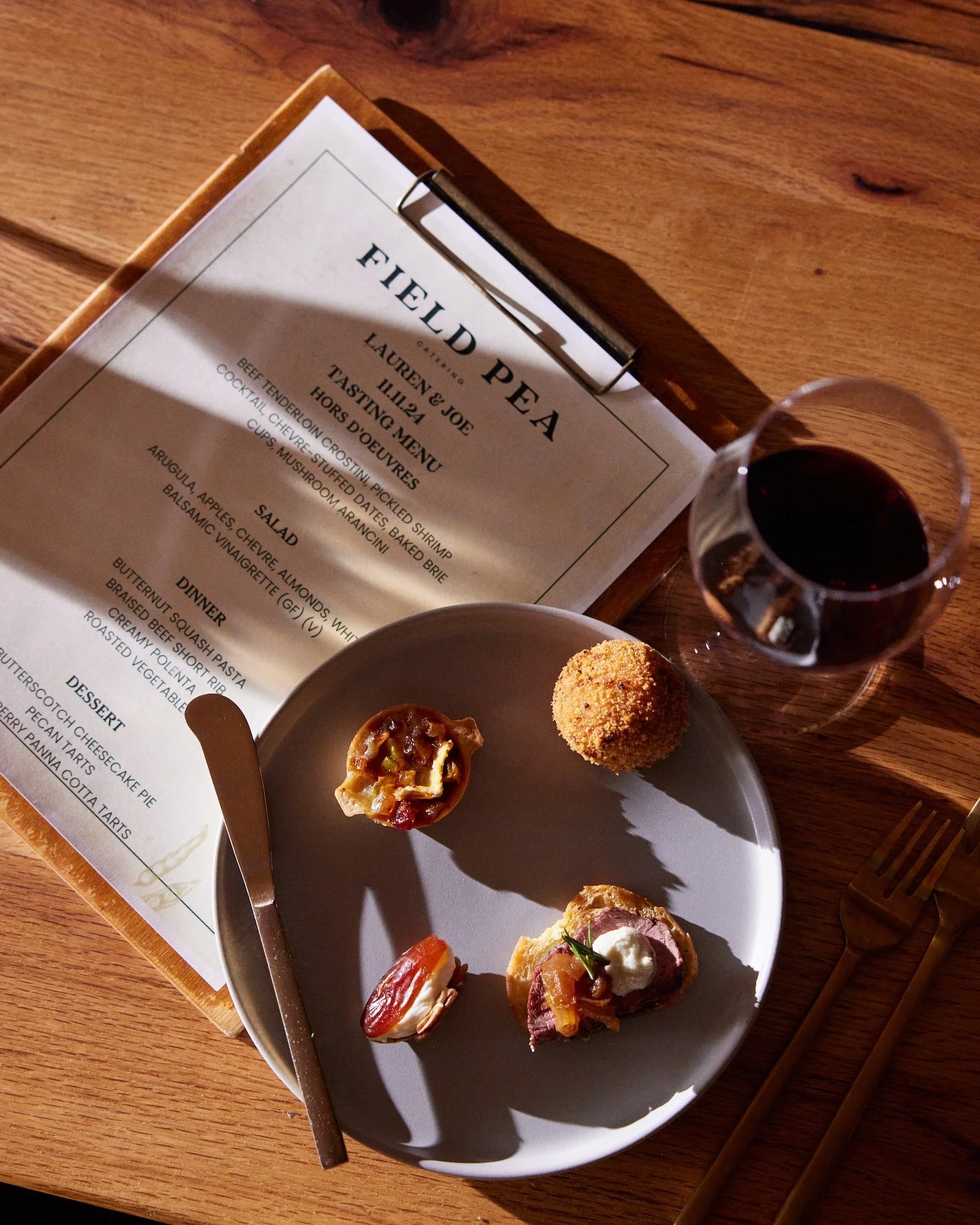 A plate with three appetizers: one with a topping, a spherical fried item, and a small open-faced roast beef sandwich with toppings. A glass of red wine, a knife, and fork are on the table. A menu and a wooden serving tray are also visible.