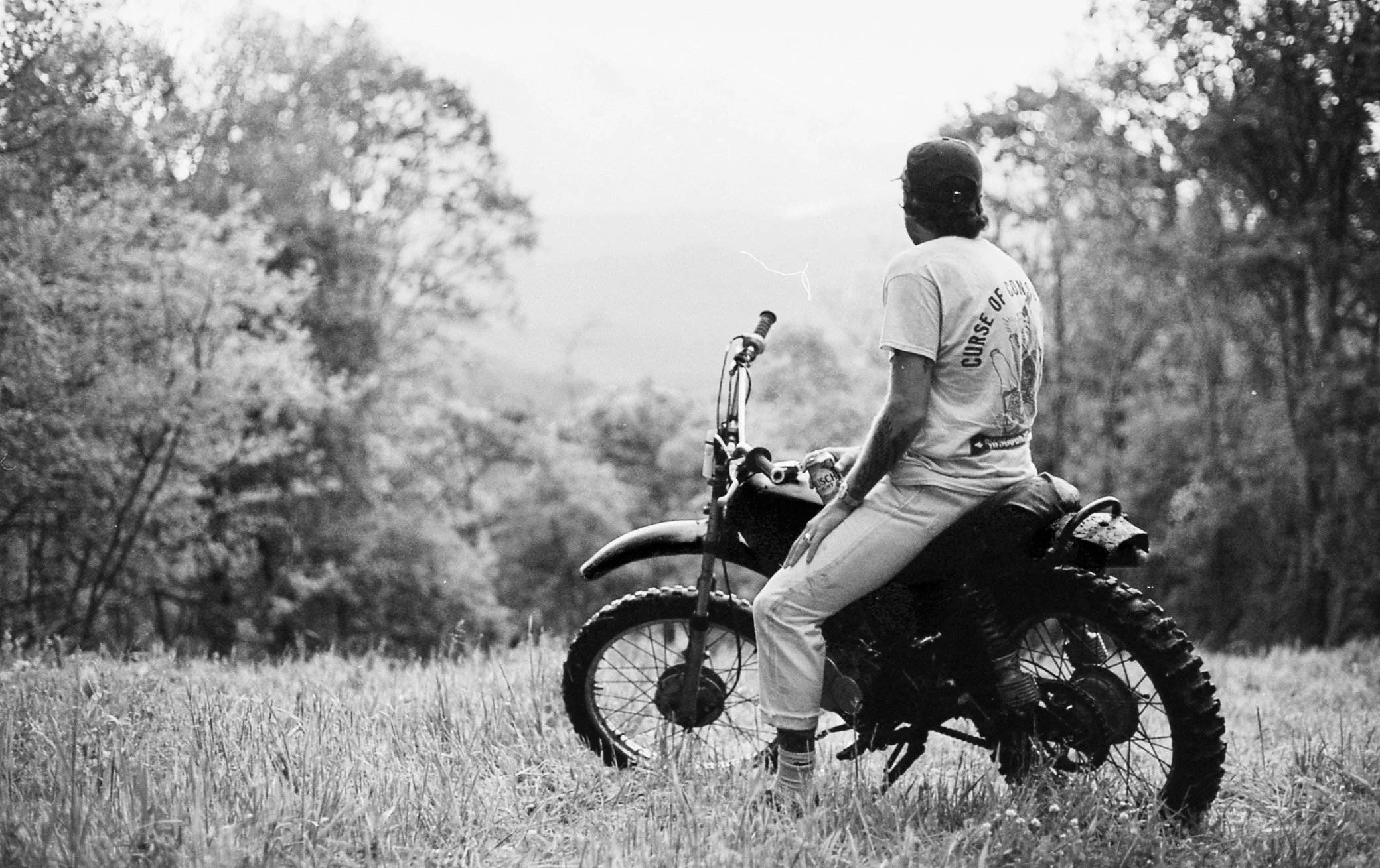 Person sitting on a dirt bike in a grassy field surrounded by trees, black and white photo.