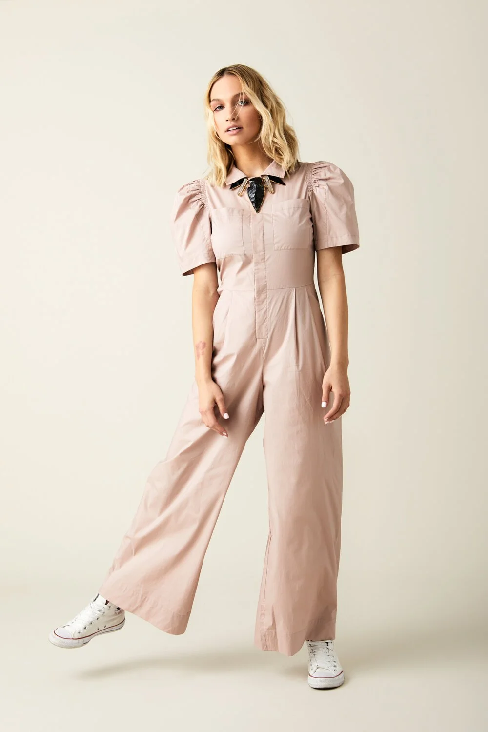 A woman in a light pink jumpsuit with puffed sleeves, white sneakers, and a black neck scarf standing against a plain cream background.