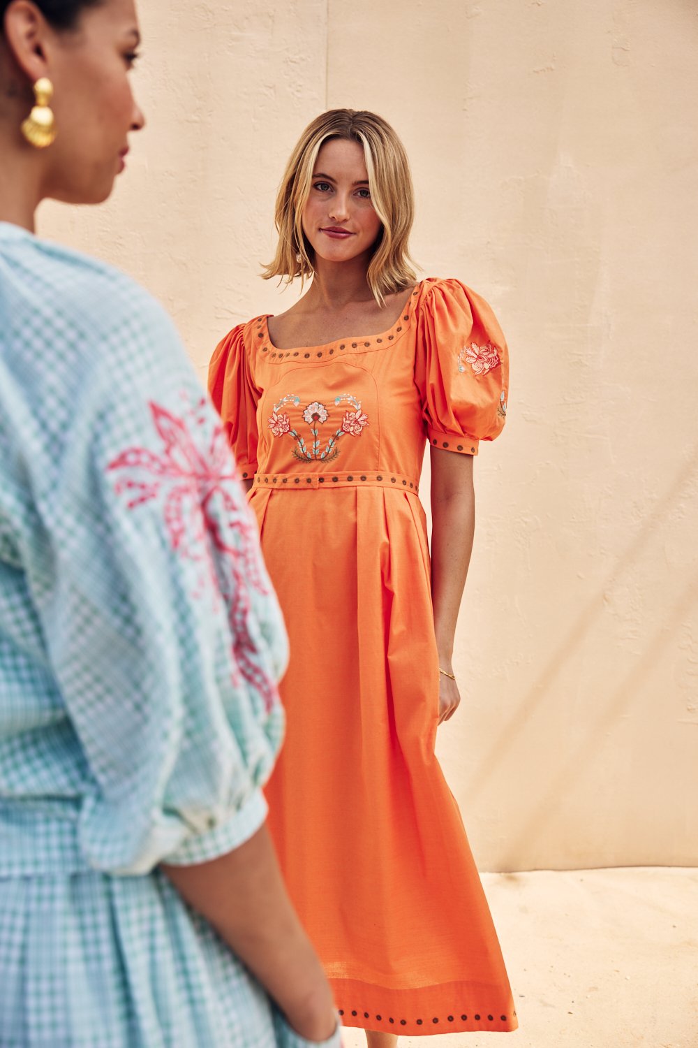 Young woman wearing an orange dress with embroidery, standing against a neutral wall, looking at another person partially in frame.