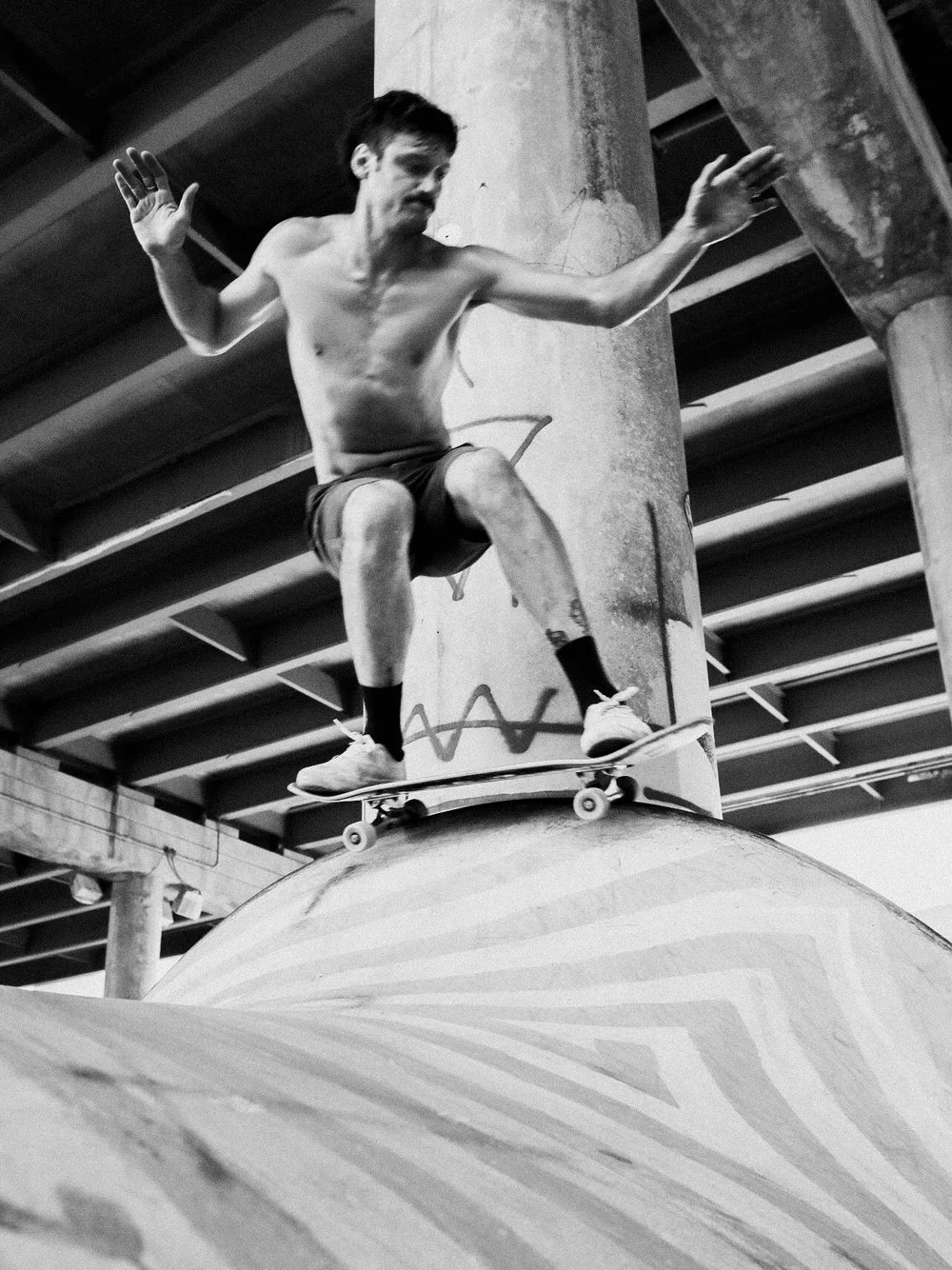 A shirtless man skateboarding on a curved ramp under a bridge or overpass in an urban setting.